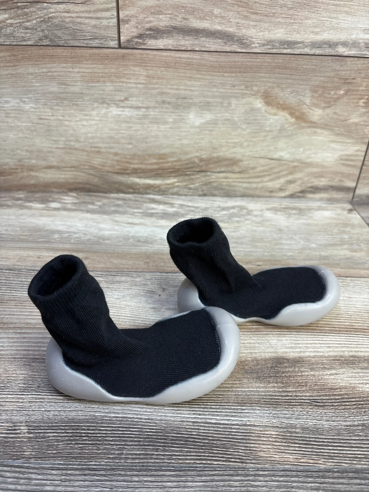 Angled view of black baby sock shoes showcasing sock-like uppers and soft rubber soles on a wood-look floor