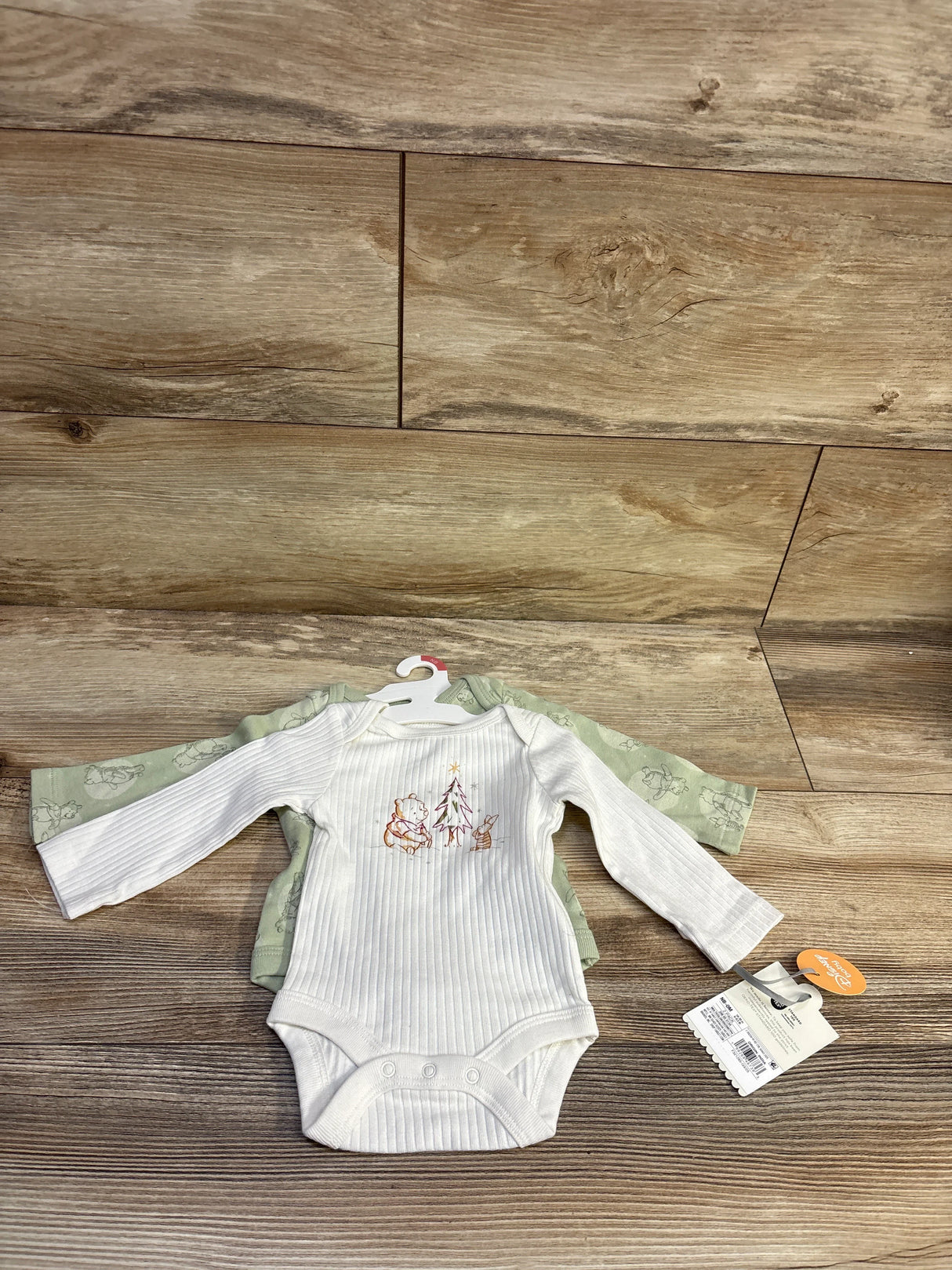 Front of Cloud Island x Disney 2-pack newborn bodysuits, ribbed white Pooh-embroidered and green printed on wood floor