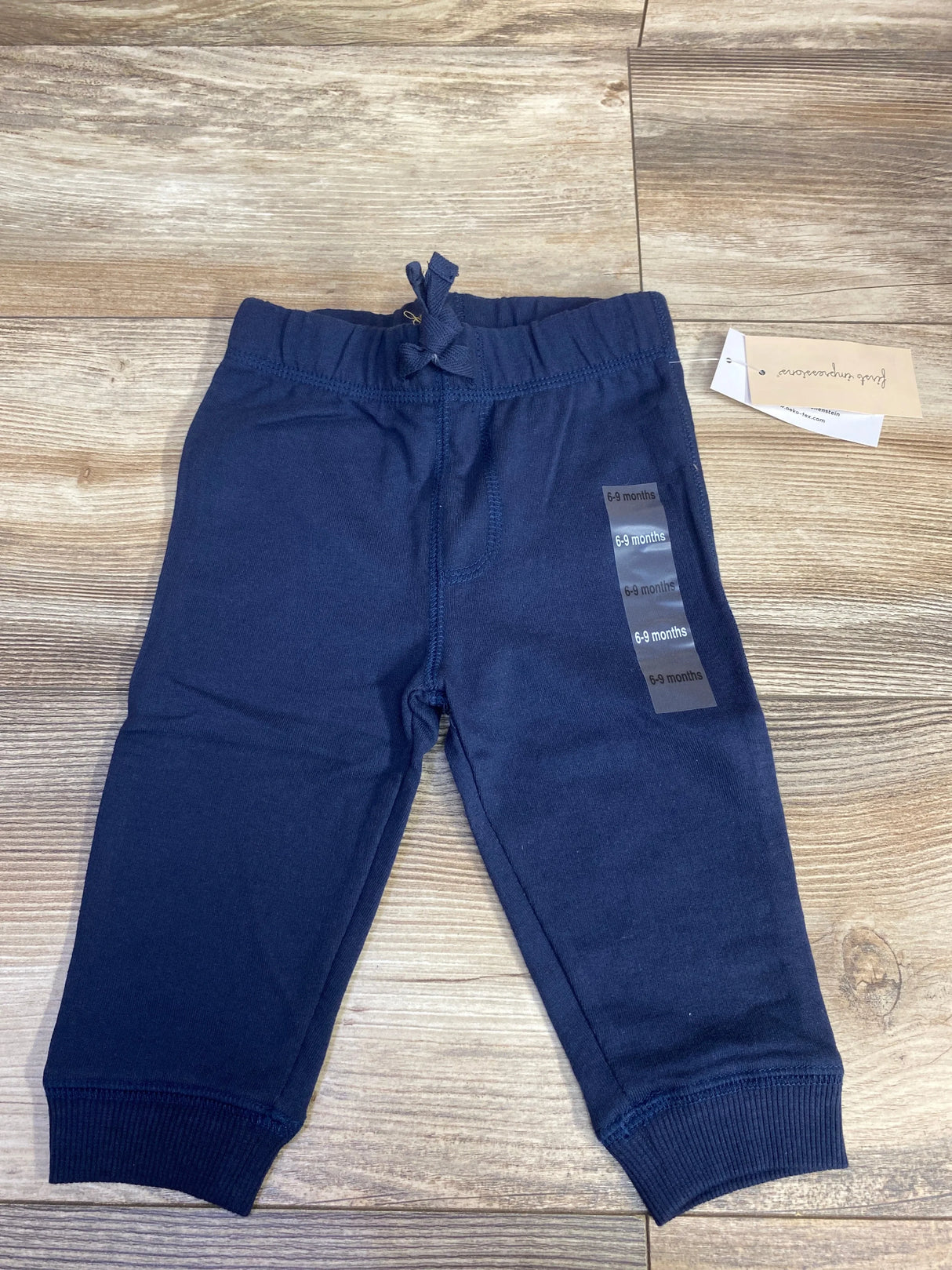 Top-down view of First Impressions navy pull-on baby pants showcasing elastic drawstring waist and ribbed ankle cuffs with wood floor background, size sticker reads 6-9 months