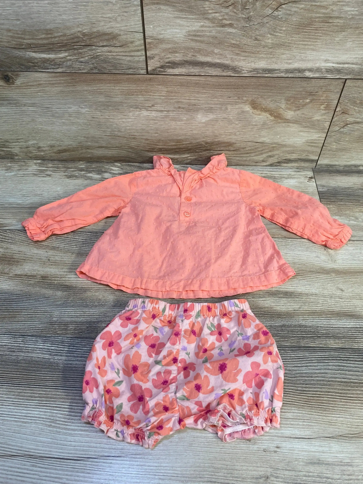 Flat lay of pink baby two-piece: swiss-dot long-sleeve shirt with ruffled collar and floral elastic-waist shorts on wood background