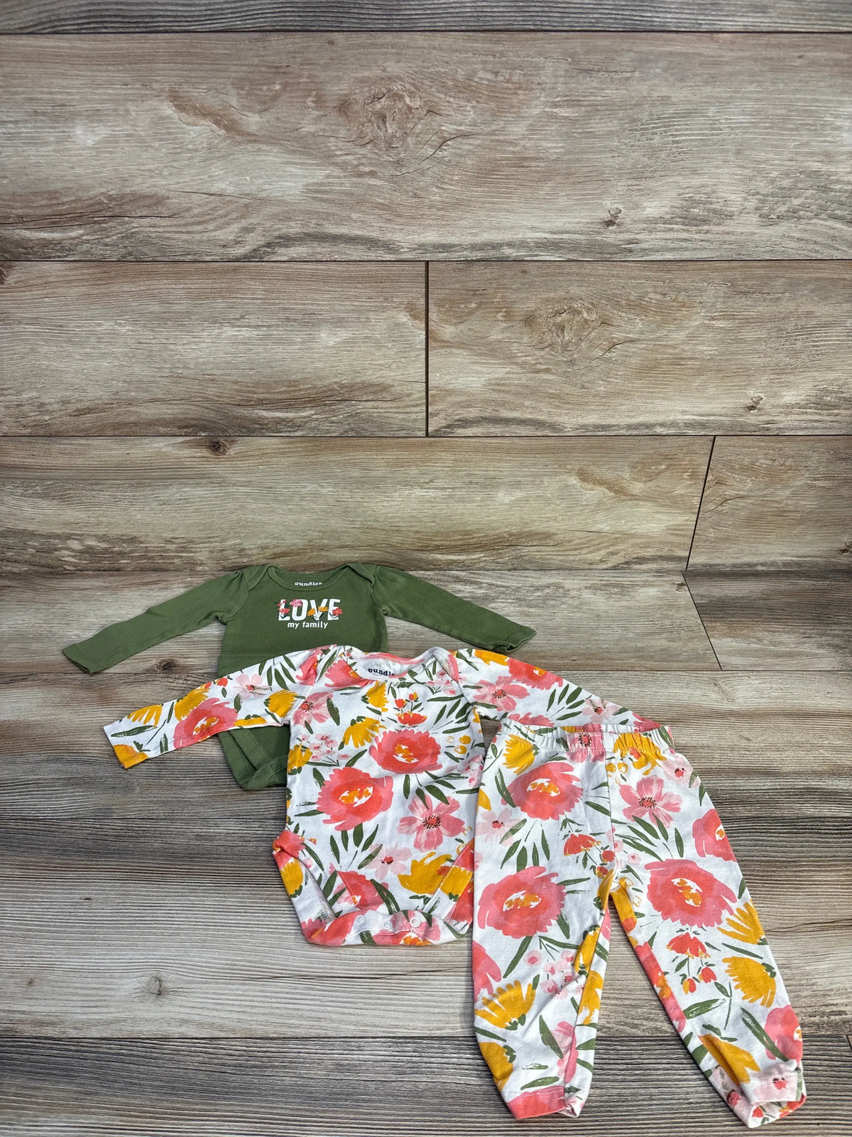 Top view of Children’s Place three-piece bodysuit and pant set showcasing floral and green garments on wood flooring