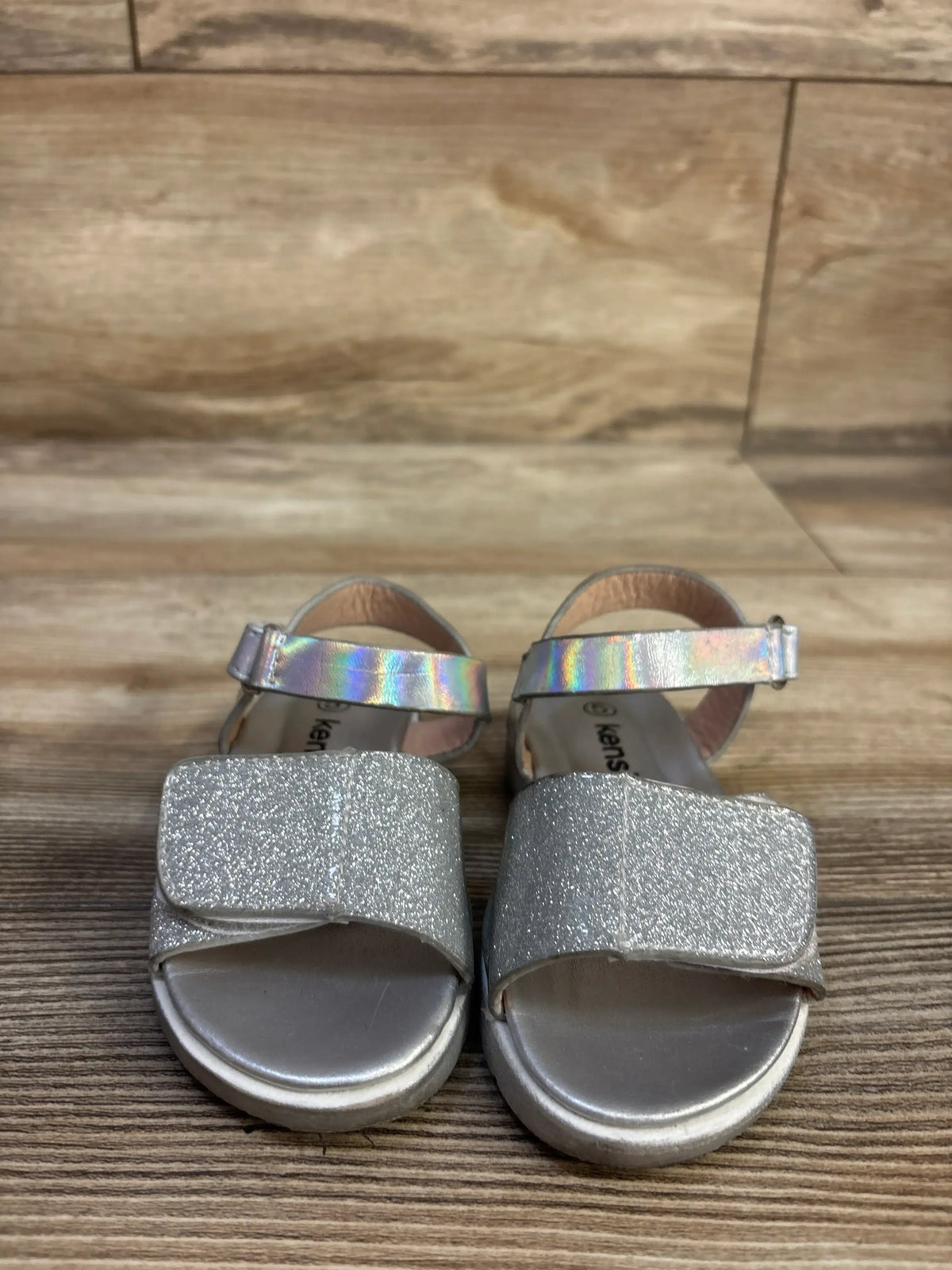 Front view of Kensie Girl silver glitter sandals showcasing wide glitter straps and ankle straps on a wood floor