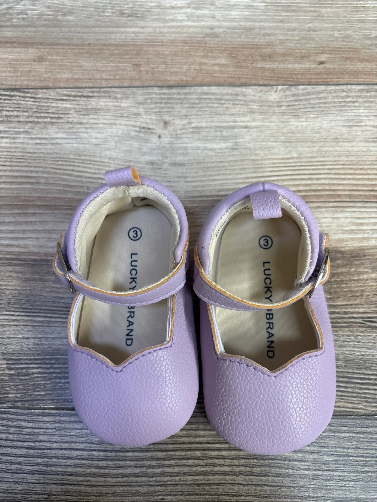 Top view of purple Lucky Brand Mary Jane crib shoes with hook and loop closures and pull tabs on wood grain
