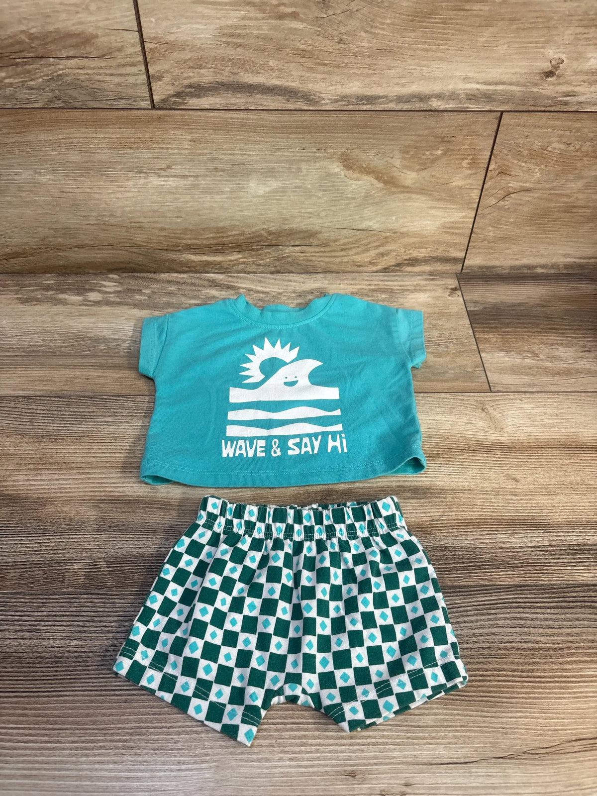 Top-down view of newborn teal shirt and shorts set showcasing Wave and Say Hi graphic and patterned shorts on wood floor