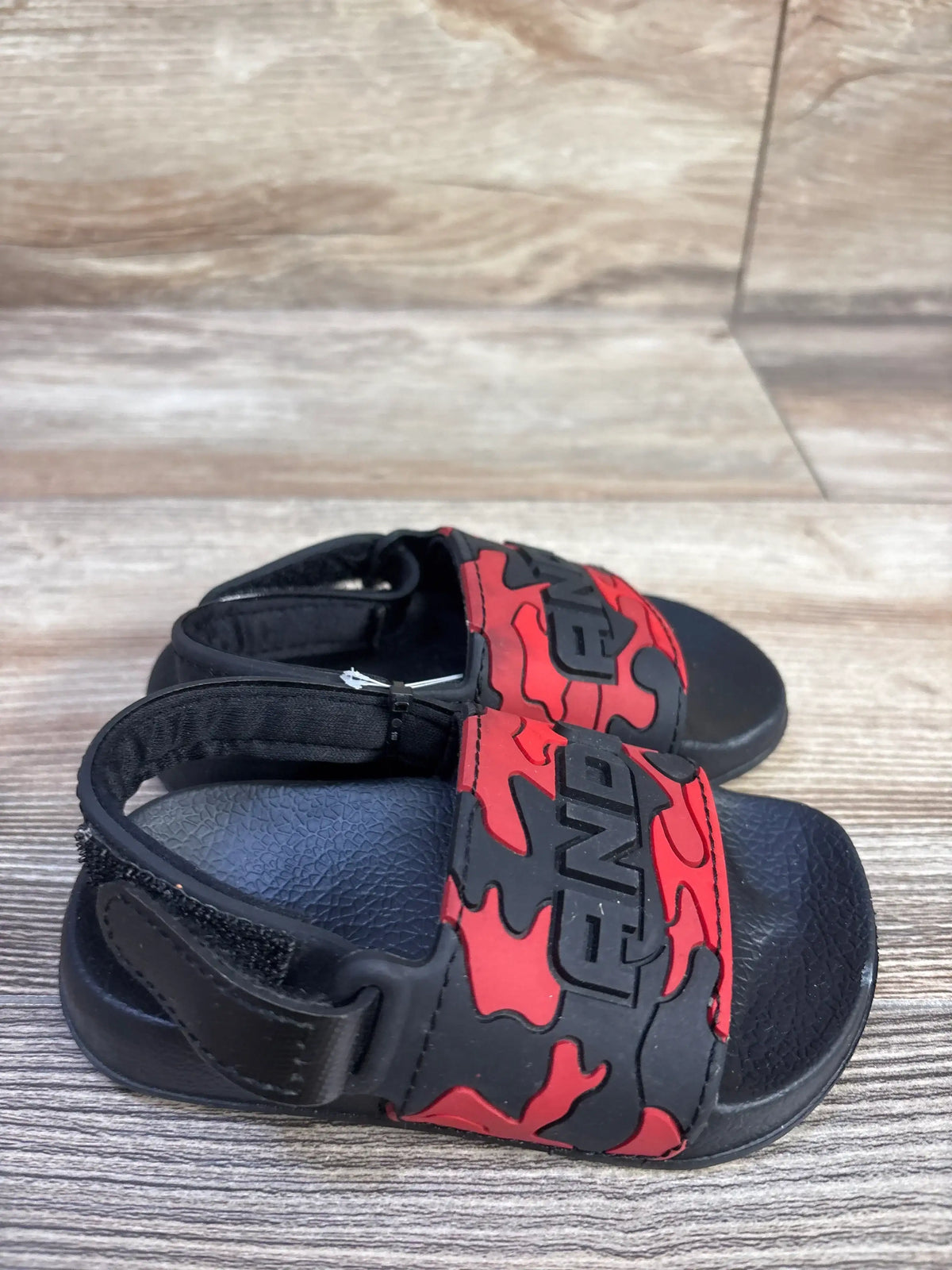 Angled front view of And1 toddler red camo slide sandals size 5-6C showing adjustable Velcro heel strap on wood floor background