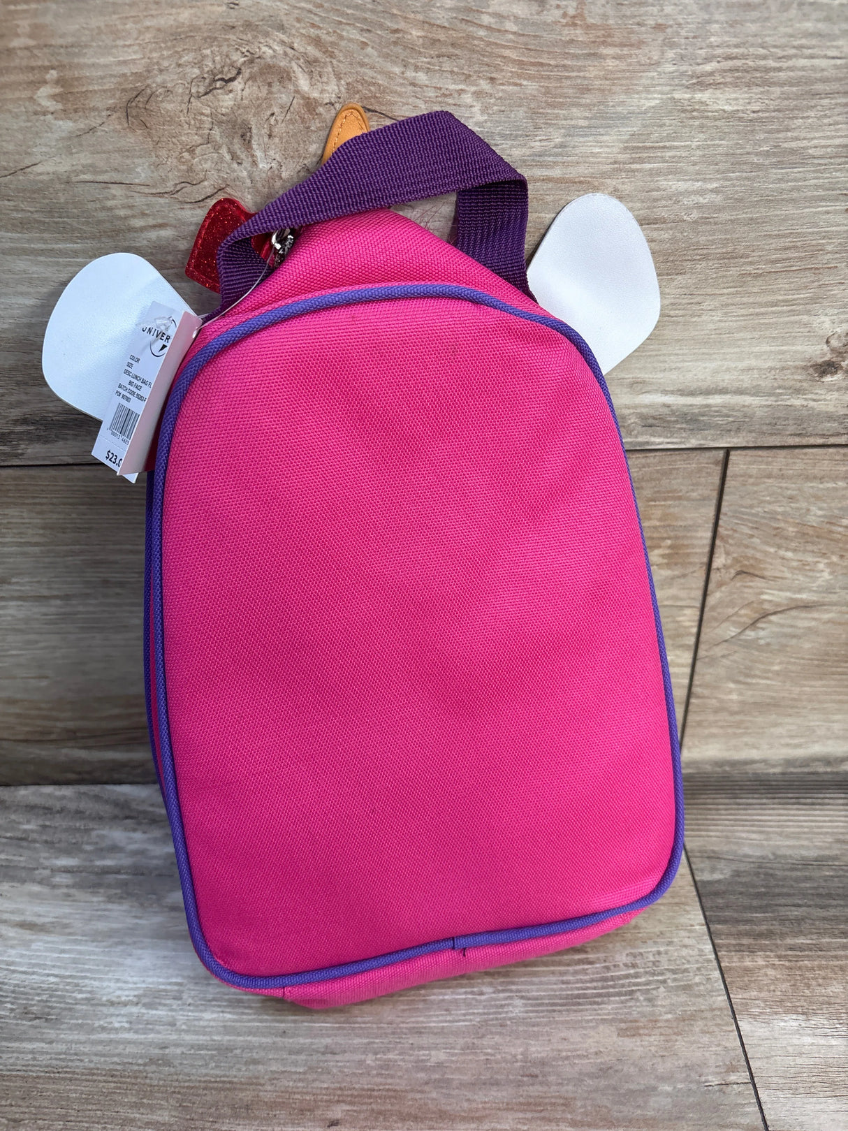 Front view of Despicable Me Fluffy Unicorn lunch bag showcasing pink body, purple trim, top handle and white unicorn ears with wood plank background