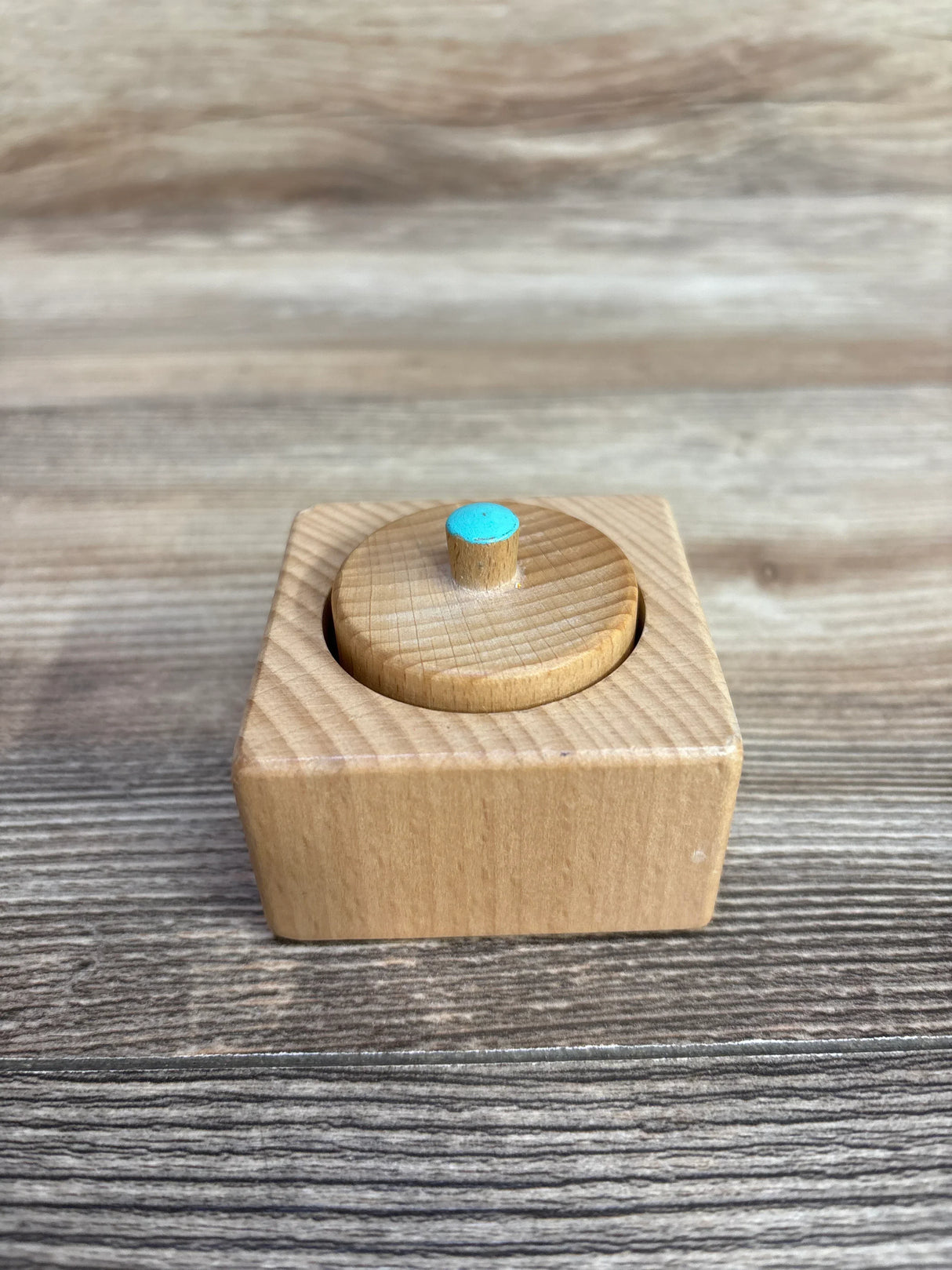 Front view of Lovevery Pincer Puzzle: square wooden box with round removable lid and blue-topped peg on wood background