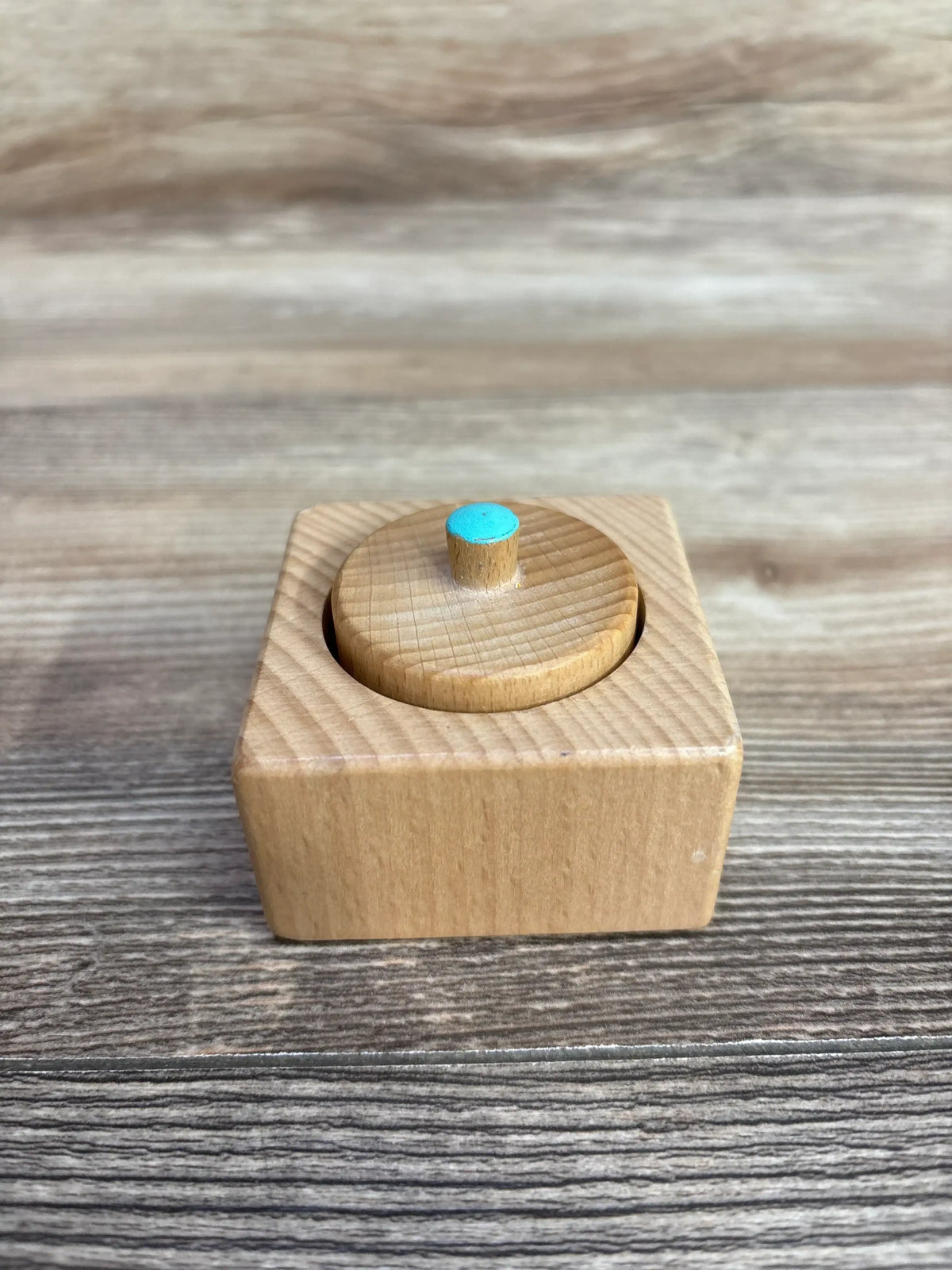 Front view of Lovevery Pincer Puzzle: square wooden box with round removable lid and blue-topped peg on wood background