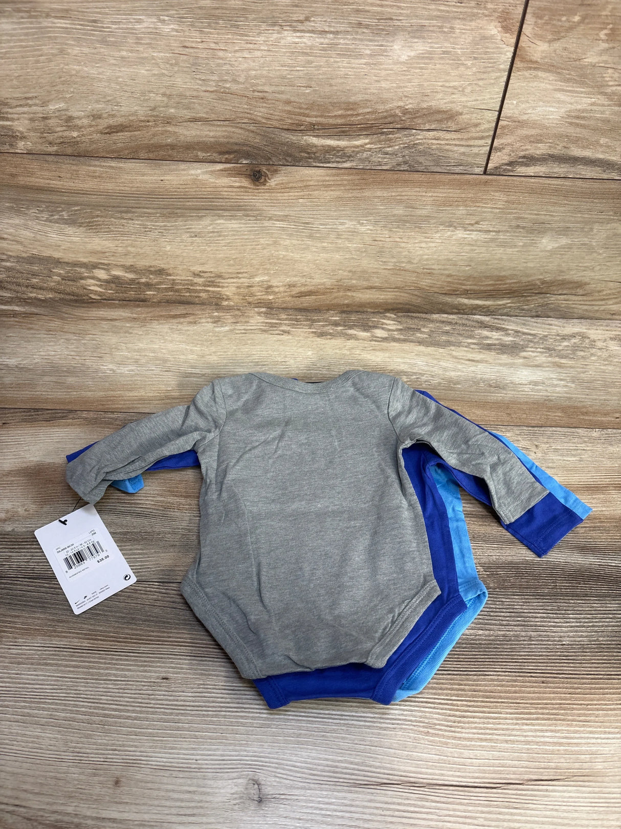 Flat lay of 3-pack long-sleeve baby bodysuits showing back of gray top and two blue bodysuits with tag on wood floor