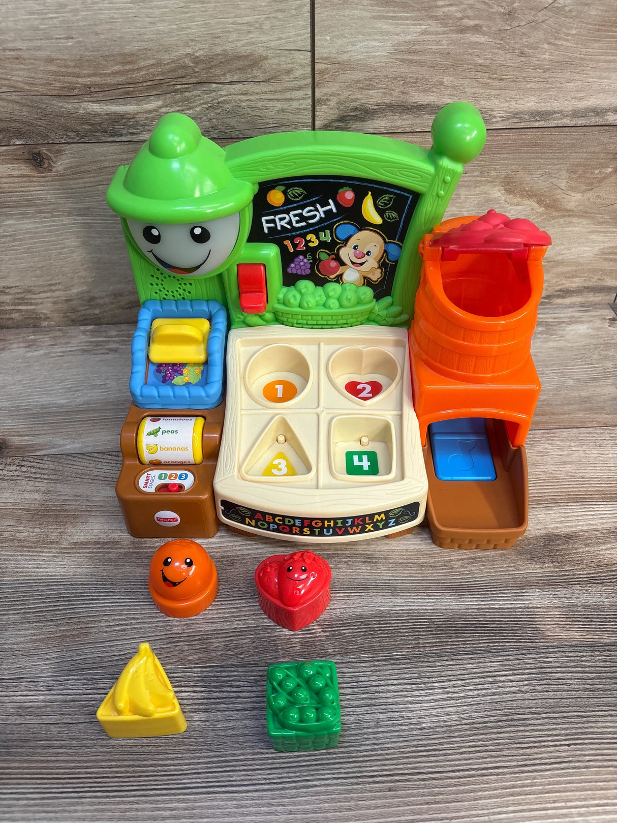Front view of Fisher-Price Laugh & Learn market showing shape-sorter playset with five fruit pieces on wood background