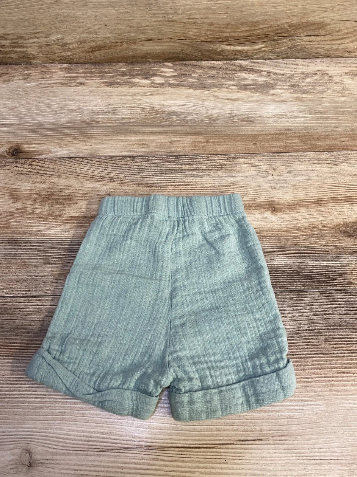 Front view of green Embrace gauze pull-on shorts showcasing elastic waistband and rolled cuffs on a wood-grain background