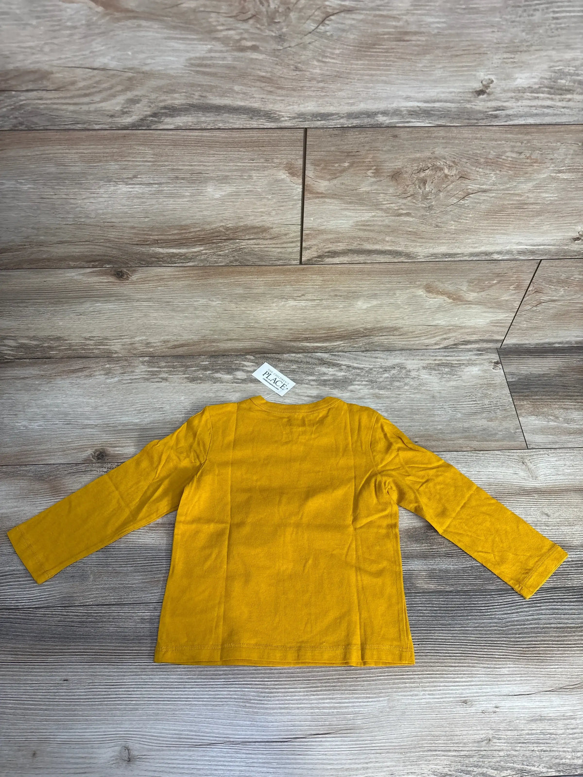 Back view of a mustard yellow long sleeve toddler shirt on wood floor with The Children's Place tag