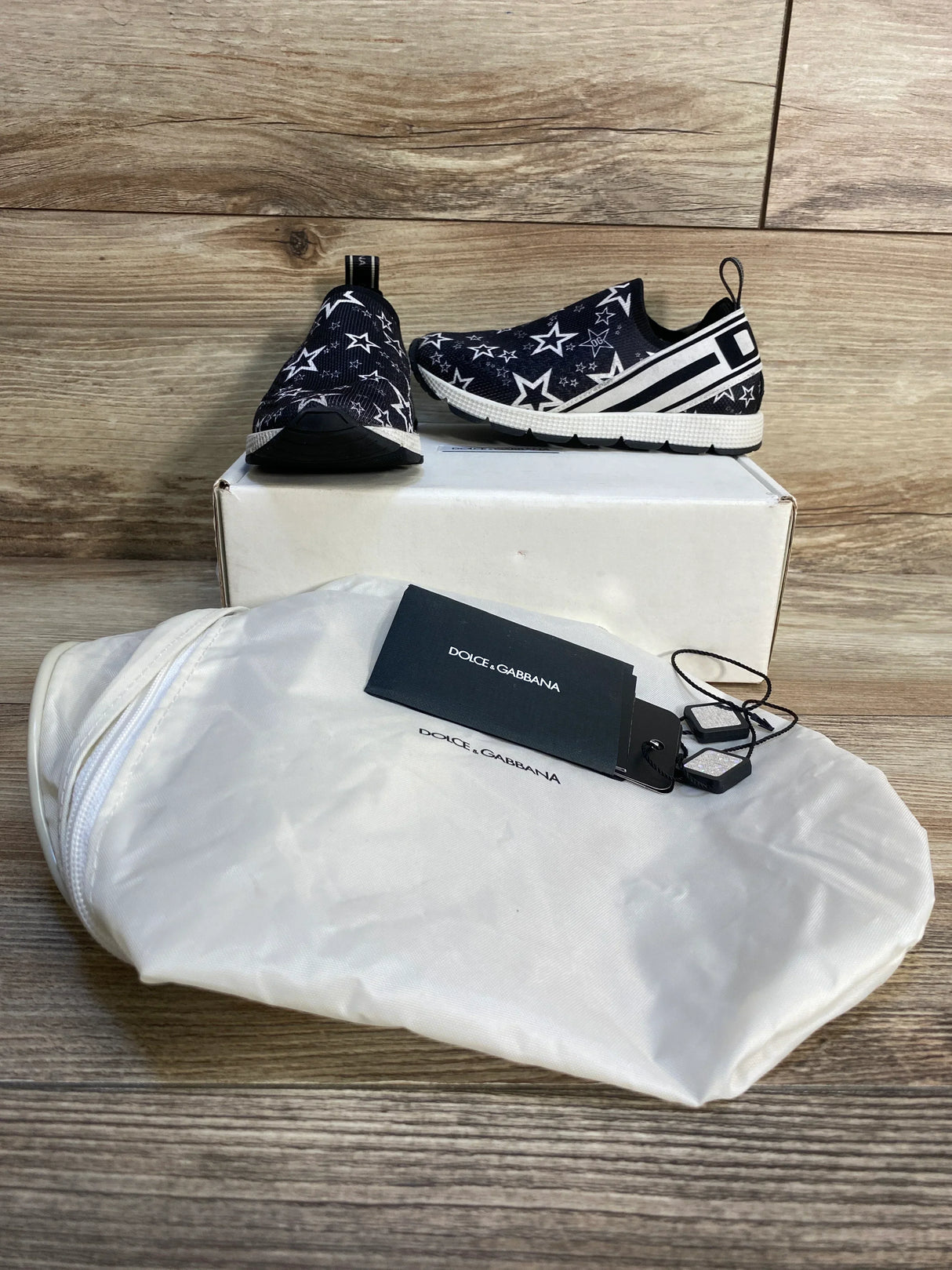Children's black slip-on sneakers with white star pattern, dust bag, and tags displayed on wood