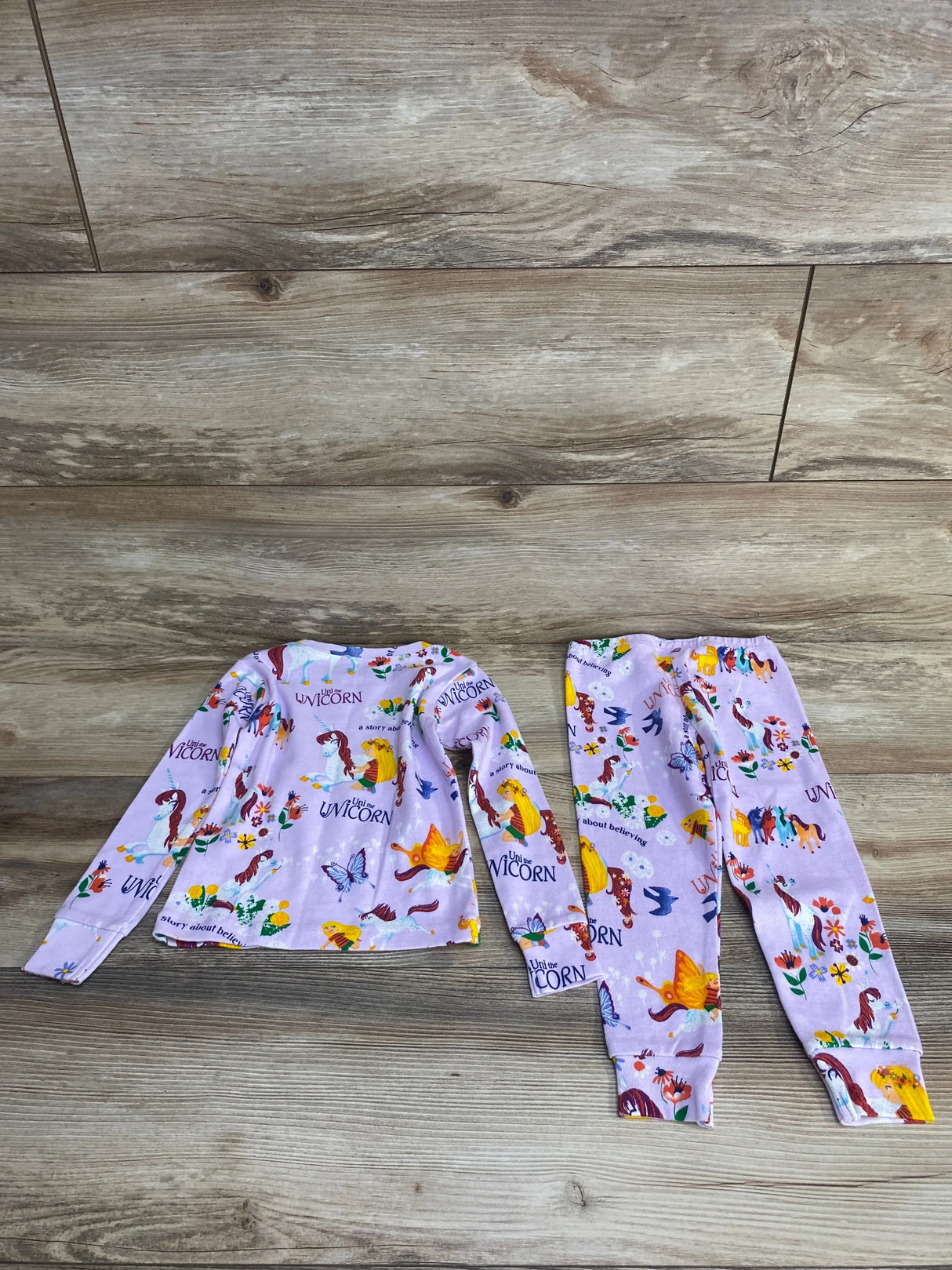 Flat lay of purple toddler Books to Bed unicorn pajama set with long-sleeve top and elastic-waist pants on wood background