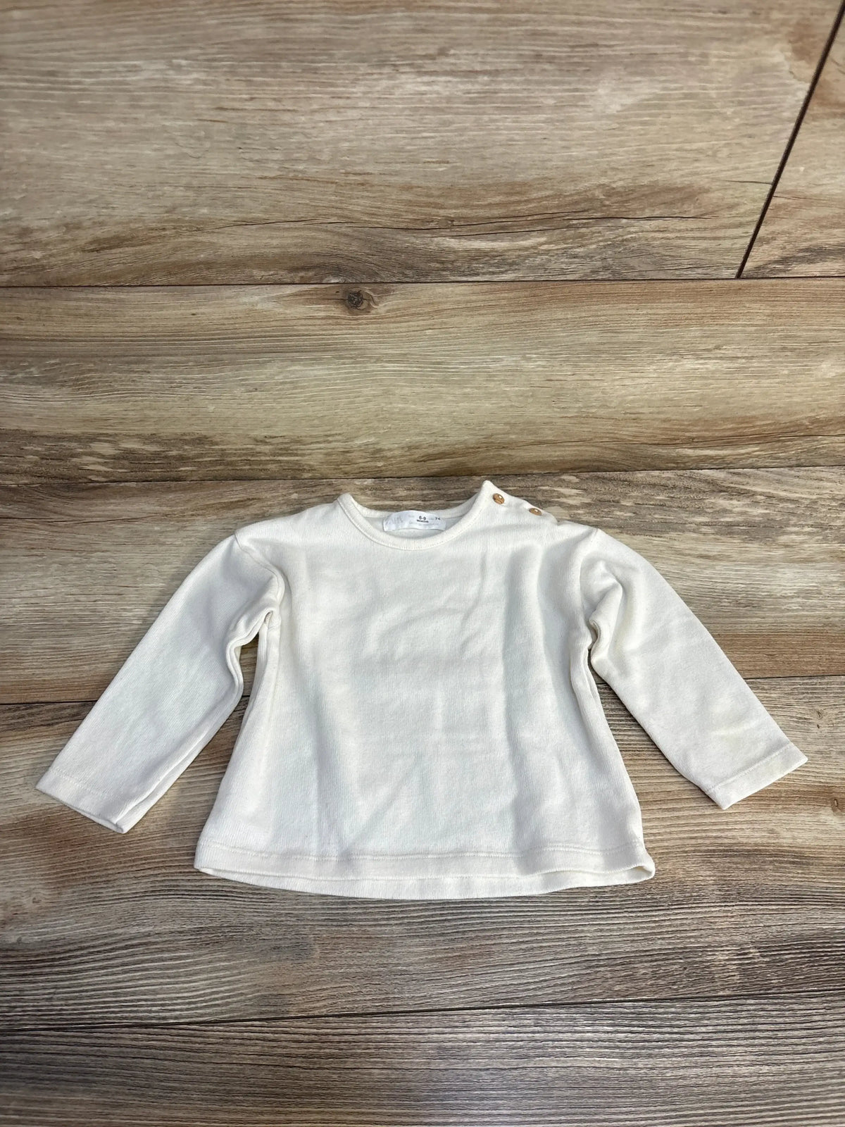 White long sleeve baby shirt with shoulder snaps, size 6-9 months, laid on wood floor