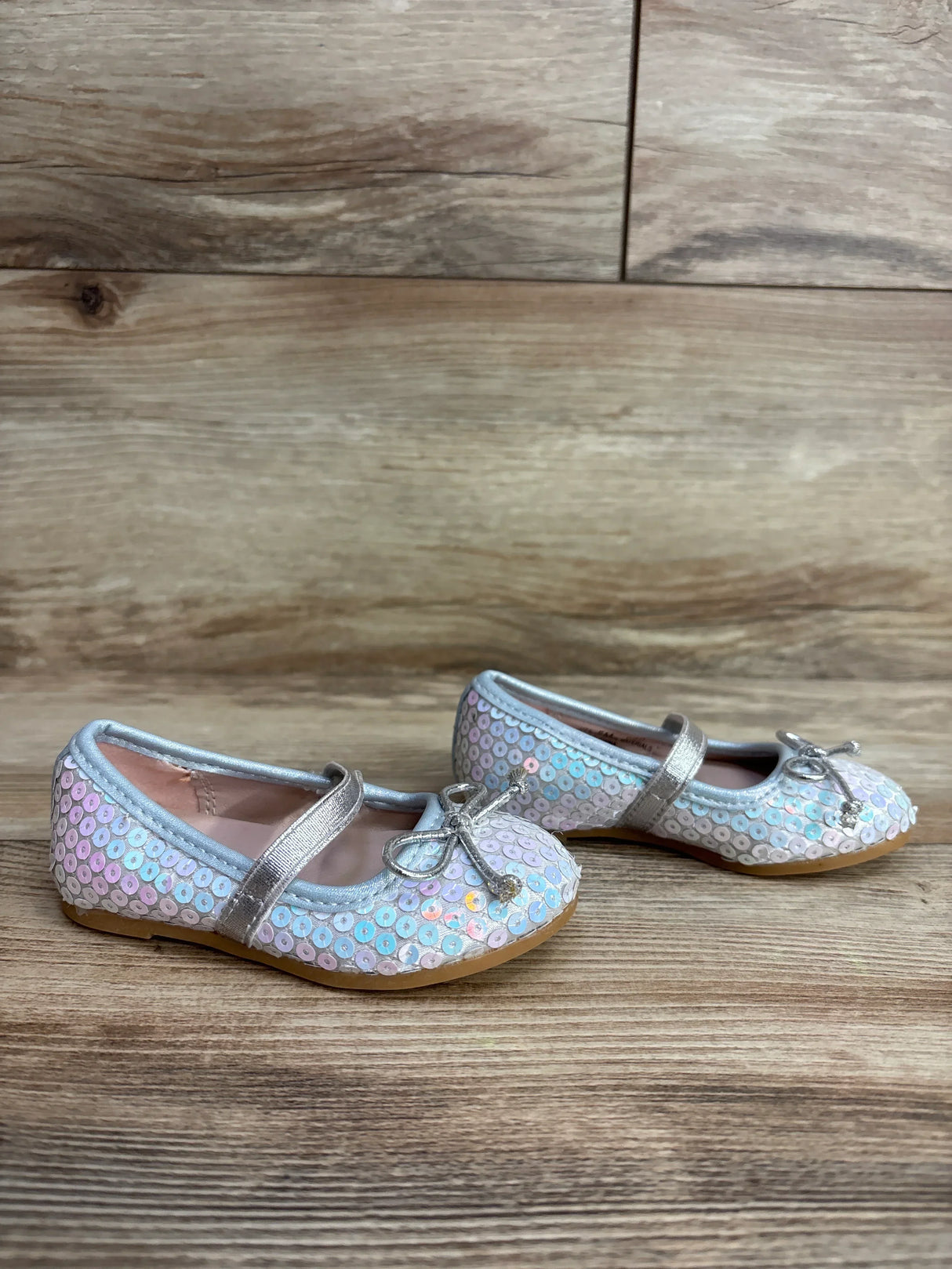 Pair of iridescent sequin toddler Mary Jane shoes with silver straps and bows on wooden floor