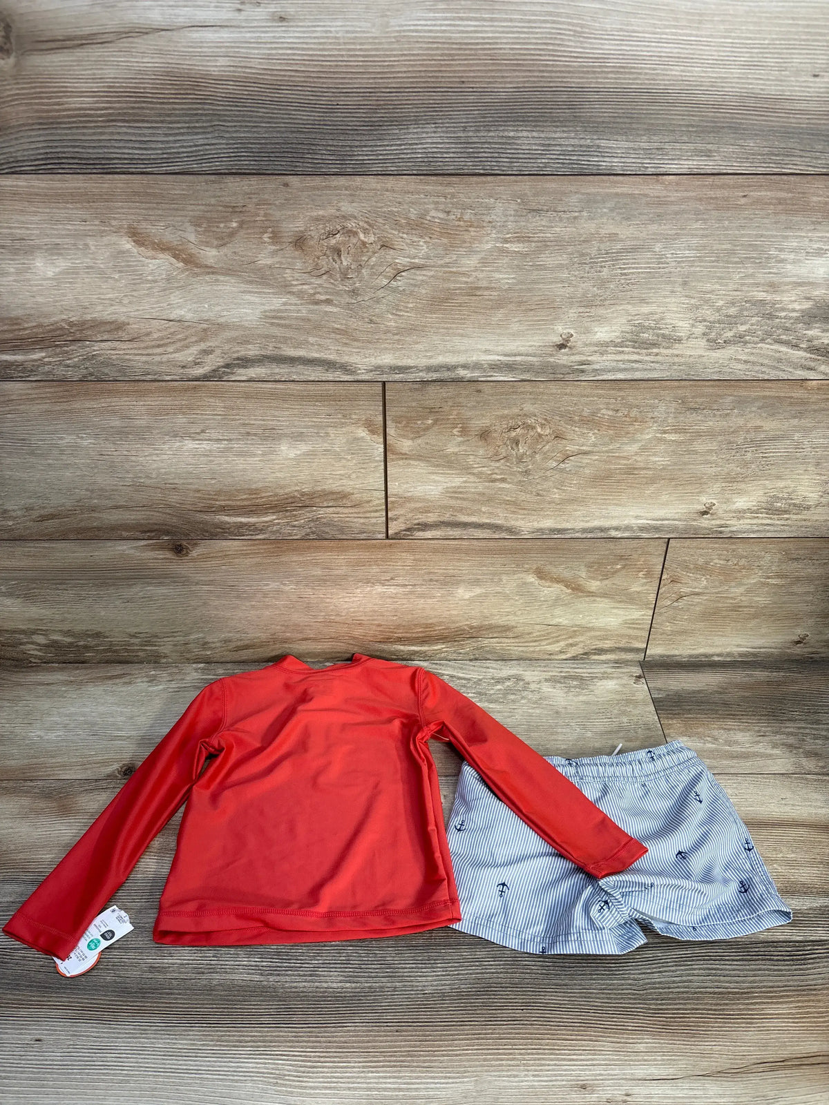 Flat lay of Just One You 2-piece rash guard set showcasing red long-sleeve top and blue striped anchor-print bottoms on wood flooring
