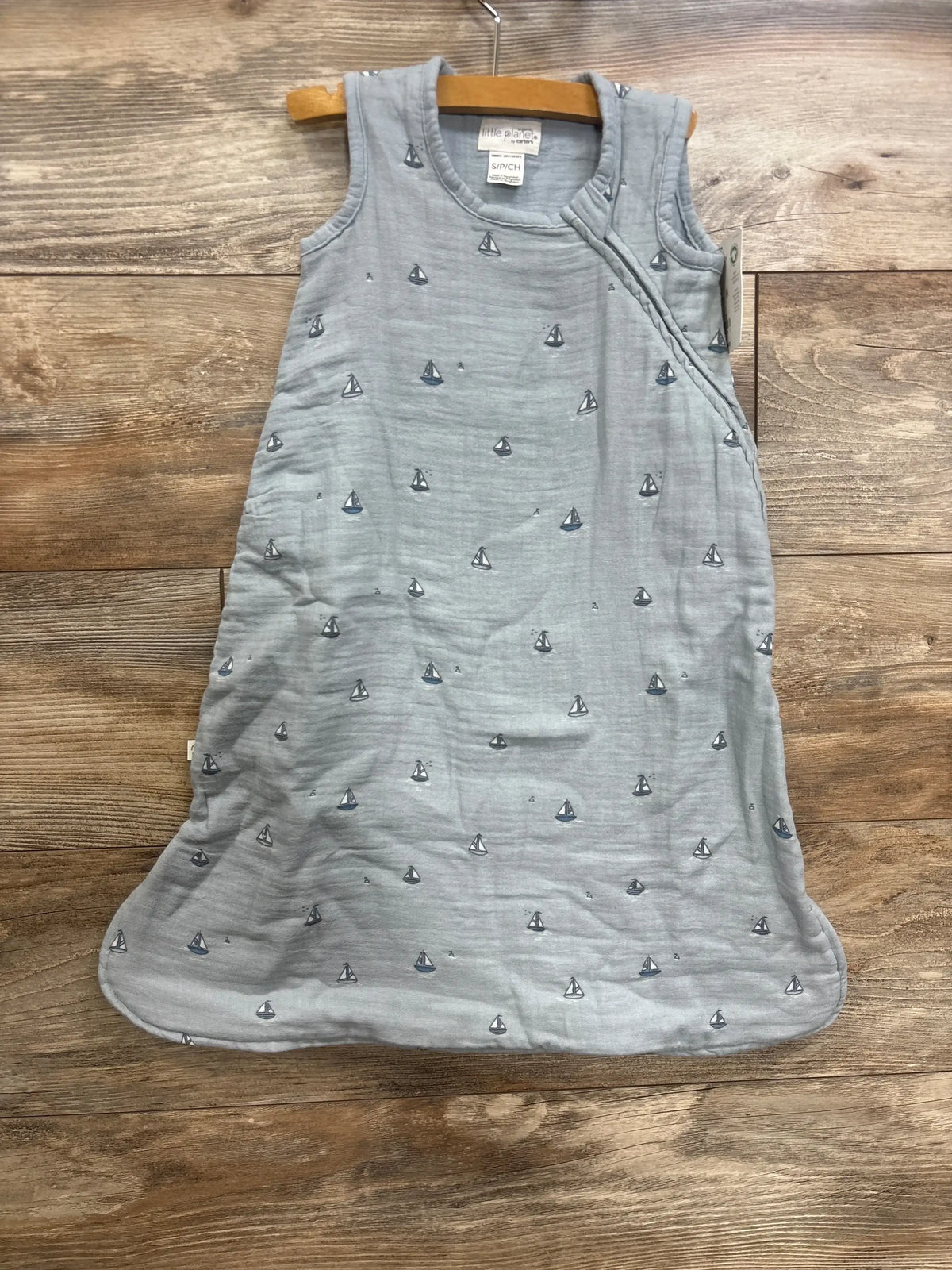 Flat lay of light blue Little Planet organic gauze sleepbag, size S 0-6m, showcasing sleeveless shape and small sailboat print on wooden floor
