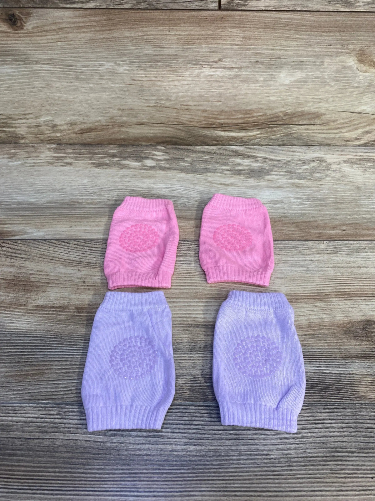 Top view of two pairs of baby crawling knee pads with textured non slip grip on pink and lilac knit, on wood background