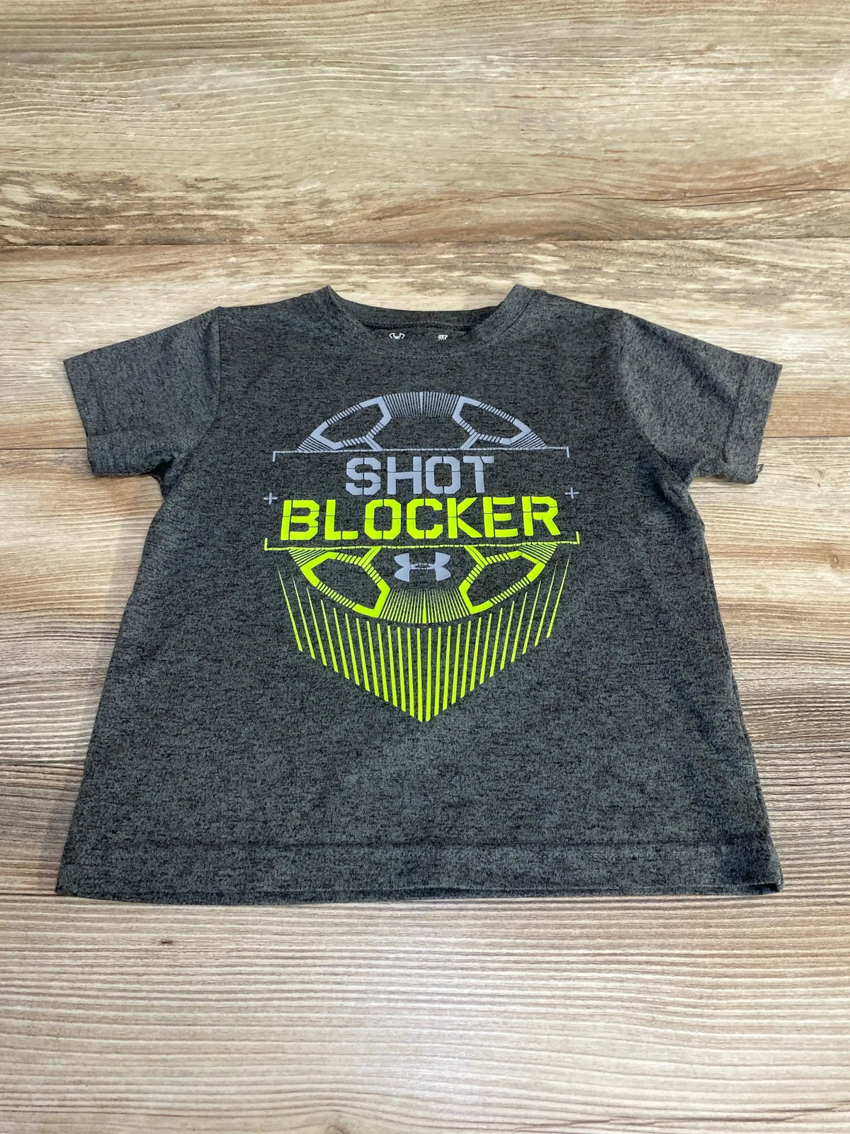 Flat lay of grey Under Armour toddler T-shirt (2T) showing the text SHOT BLOCKER in bright yellow on a light wood background