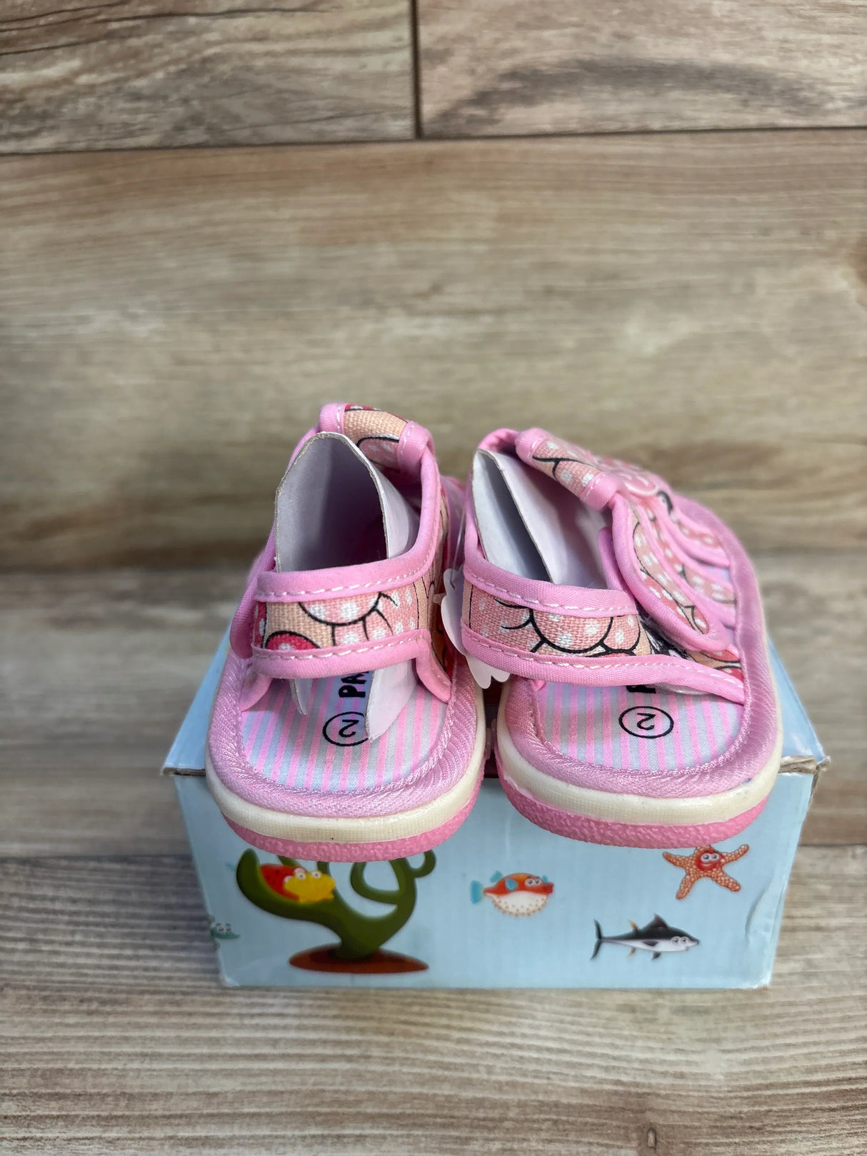Pink toddler sandals with cartoon face, displayed on a box with sea creature designs