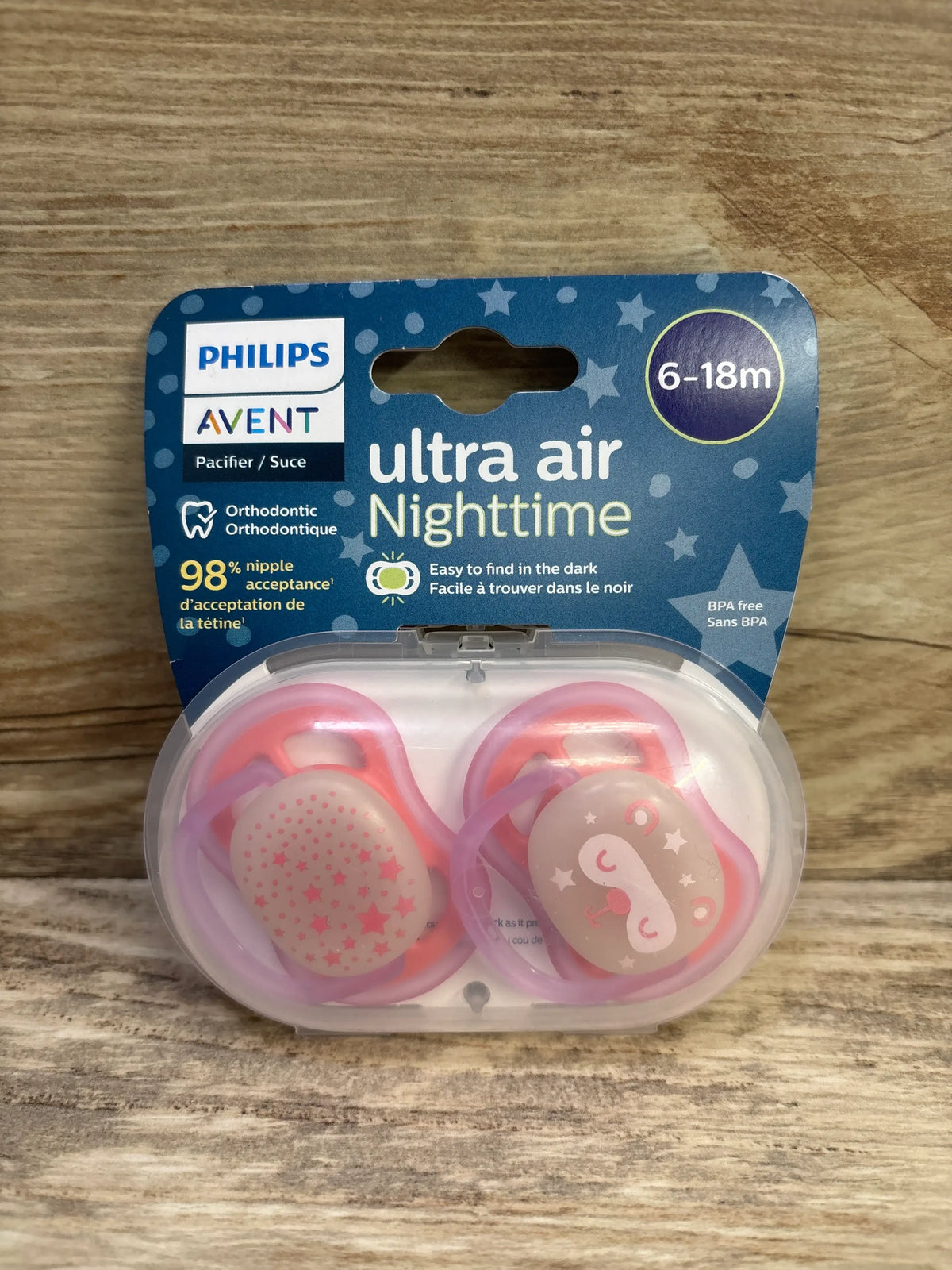 Front view of Philips Avent Ultra-Air Nighttime pink two-pack pacifiers for ages 6 to 18 months in a clear case on wood