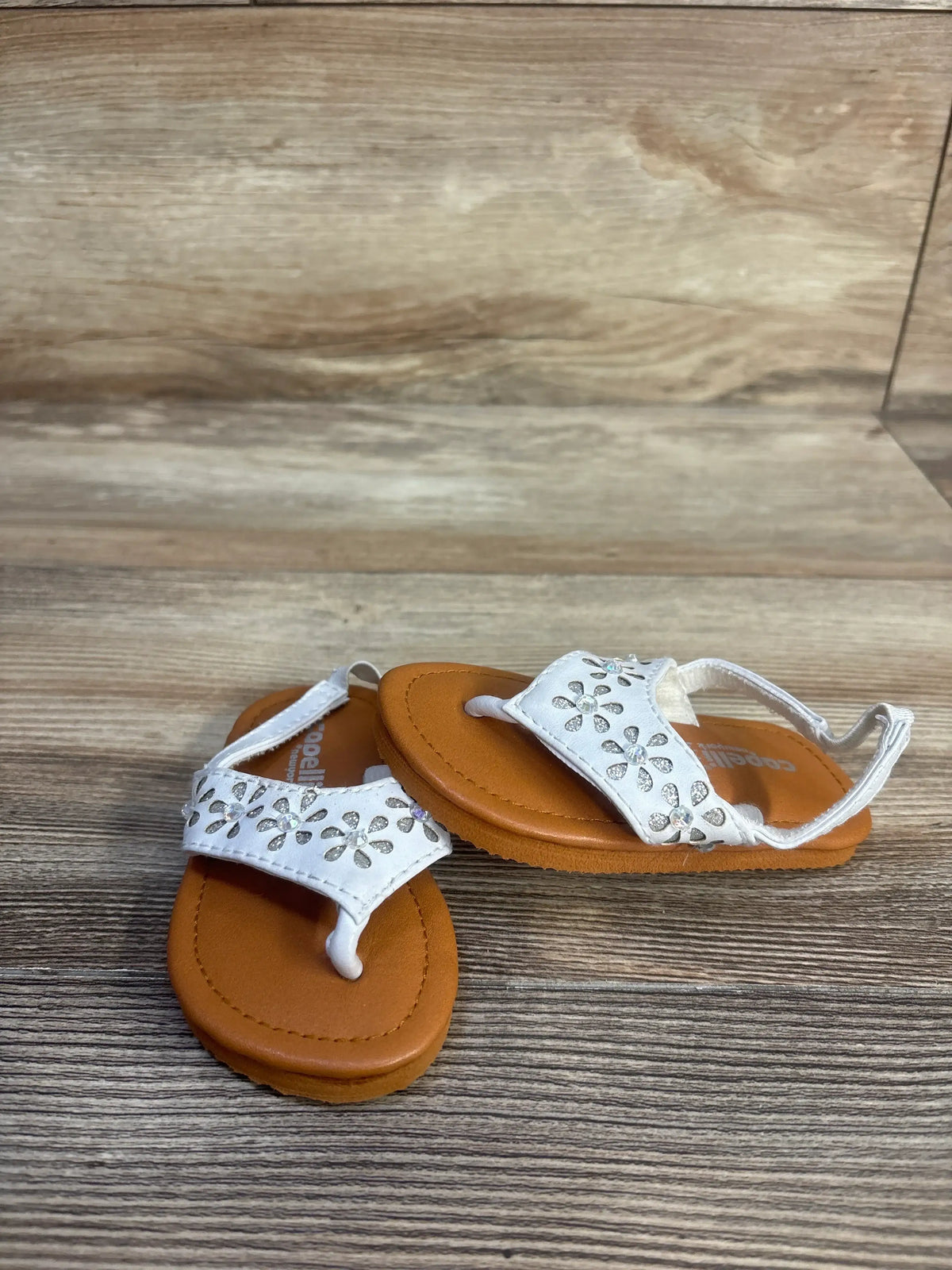 Angled view of Capelli white backstrap sandals showcasing silver floral accents and brown footbeds on a wood-grain floor