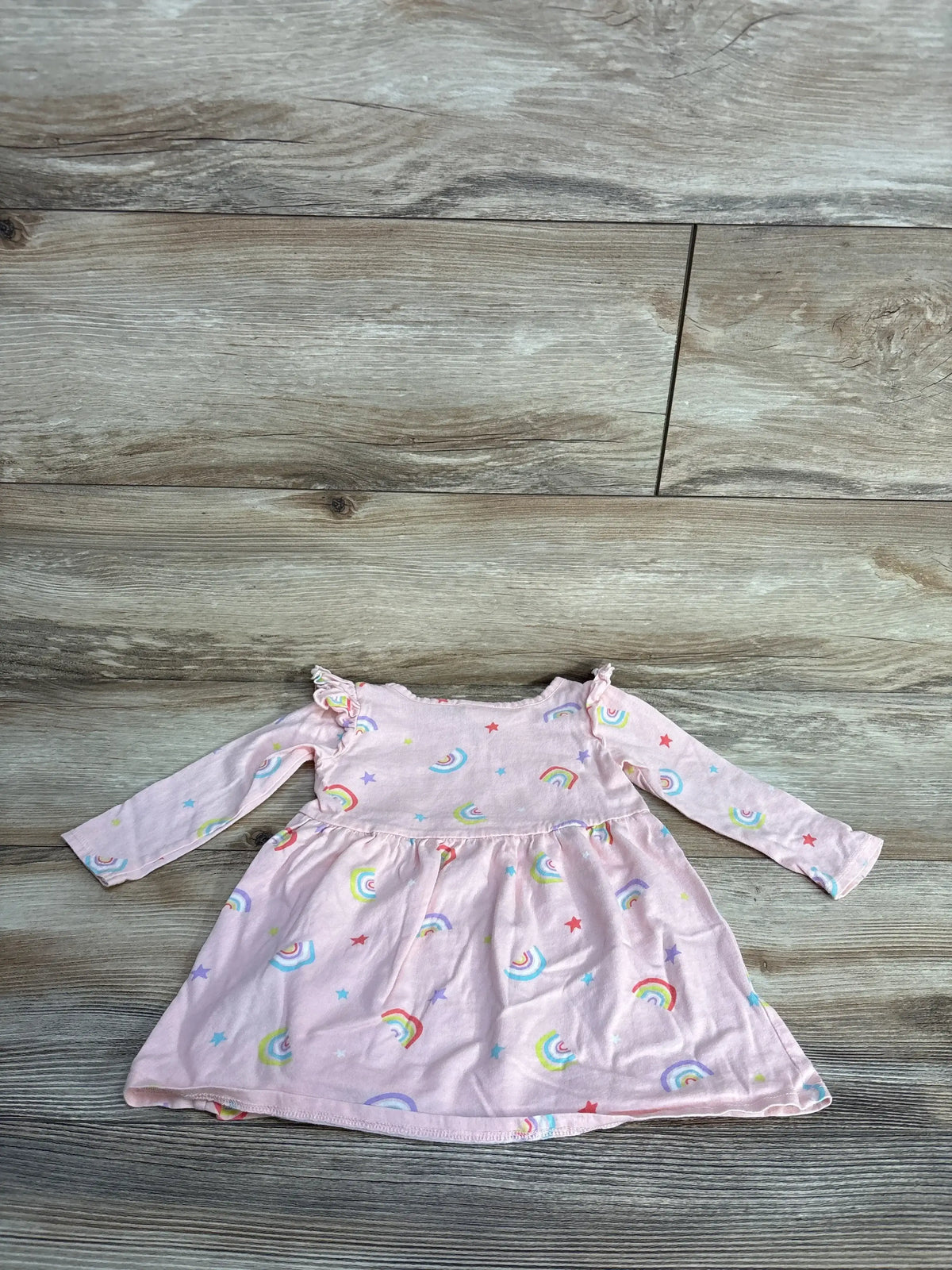Pink baby dress with rainbow and star prints, long sleeves, and ruffled shoulders on wooden floor