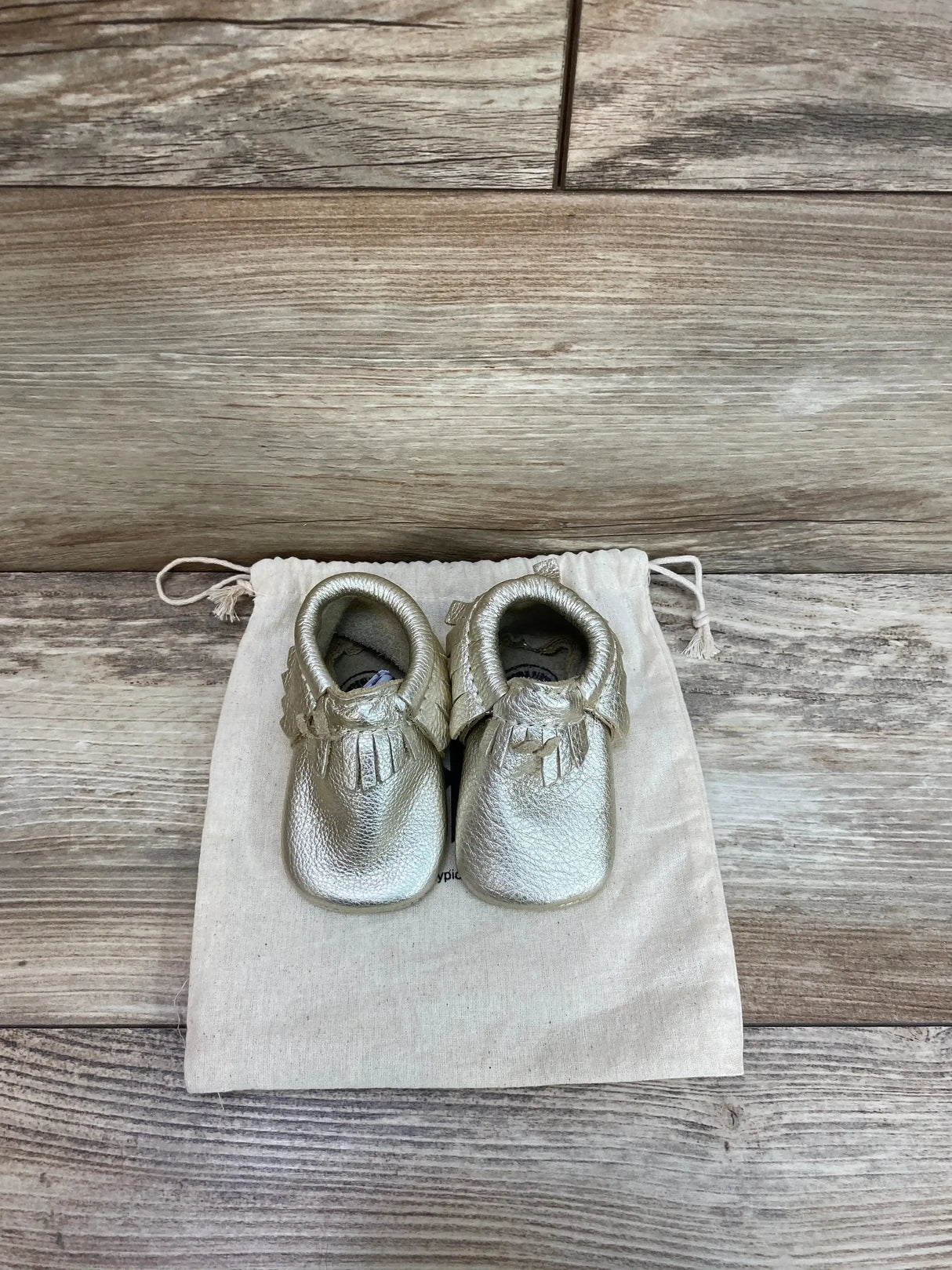 Top down view of Freshly Picked platinum baby moccasins size 2 with fringe trim on a canvas pouch and wooden floor