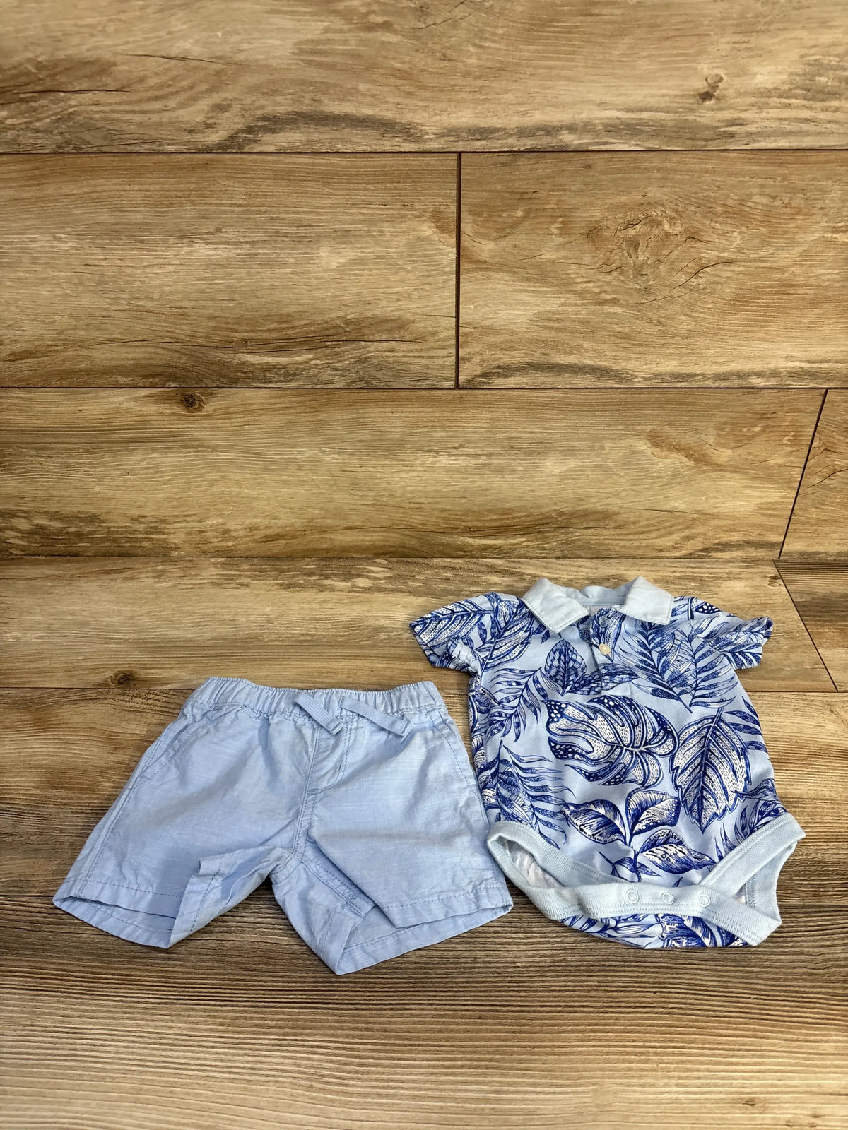 Flat lay of Children’s Place blue polo bodysuit and shorts set showcasing leaf print and collared bodysuit on wood flooring
