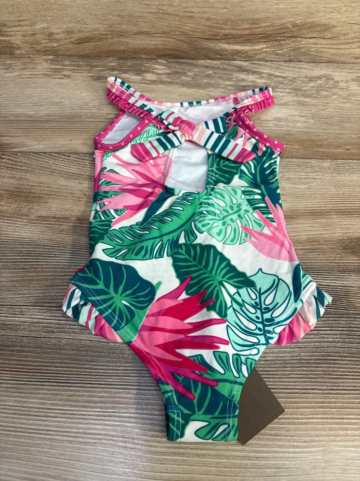 Flat lay of toddler one-piece swimsuit (size 2T) showcasing pink and green tropical leaf print, ruffle leg trim and crisscross back on light wood background