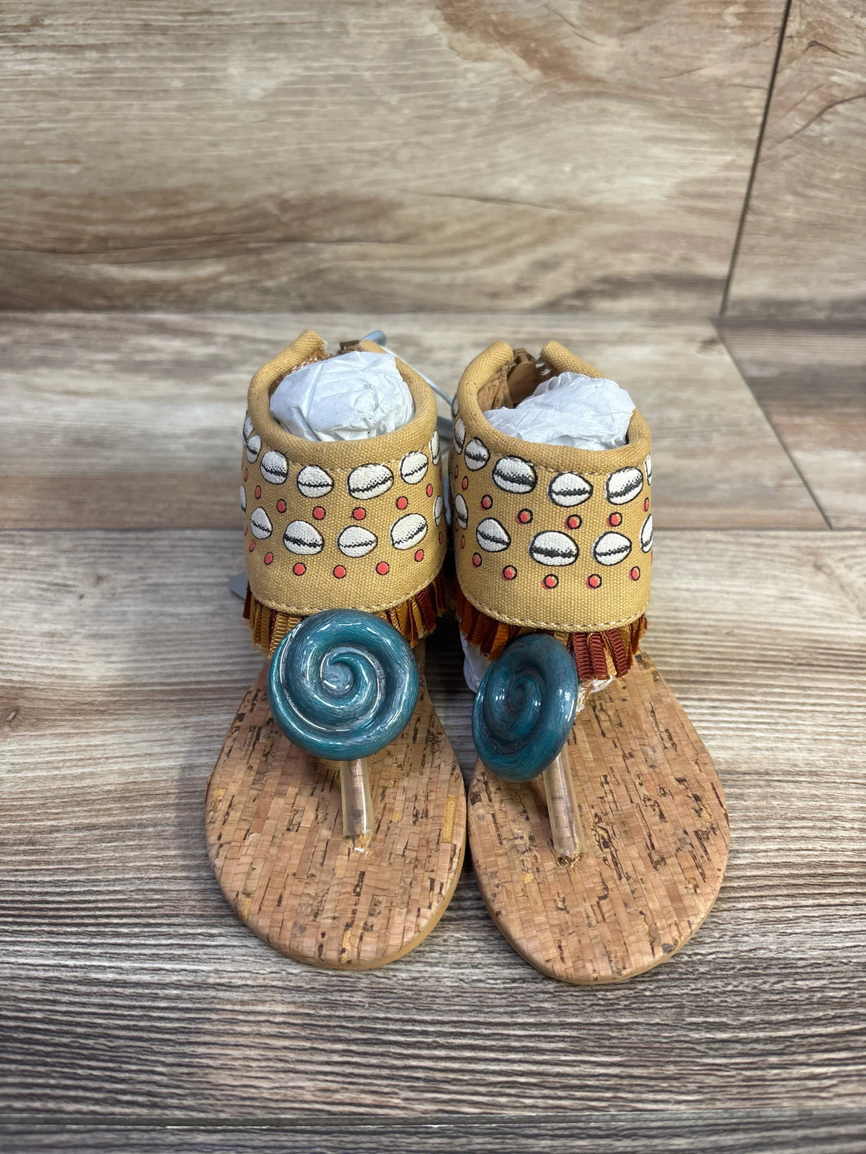 Front view of Moana toddler sandals size 5/6C with teal spiral toe charms, cork sole and yellow patterned ankle cuffs on wood