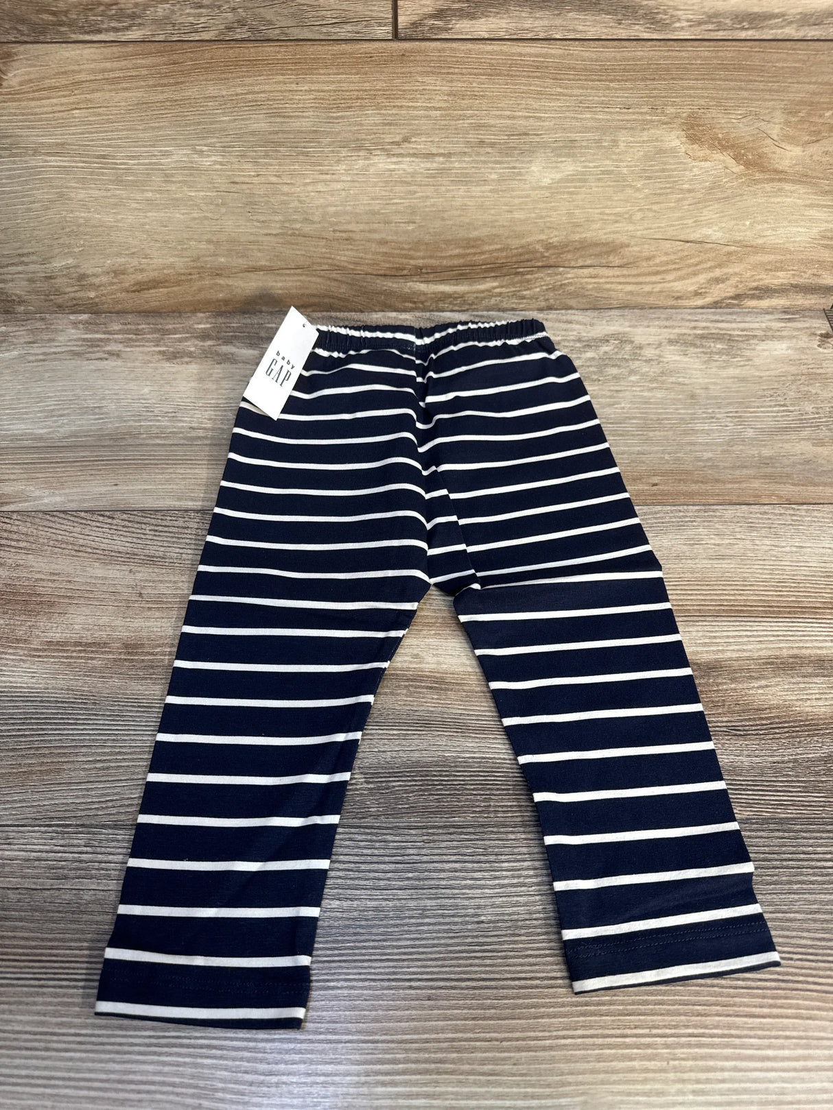 Top-down flat lay of Baby Gap navy and white striped leggings (12–18 months) showing elastic waistband and tag on wood floor