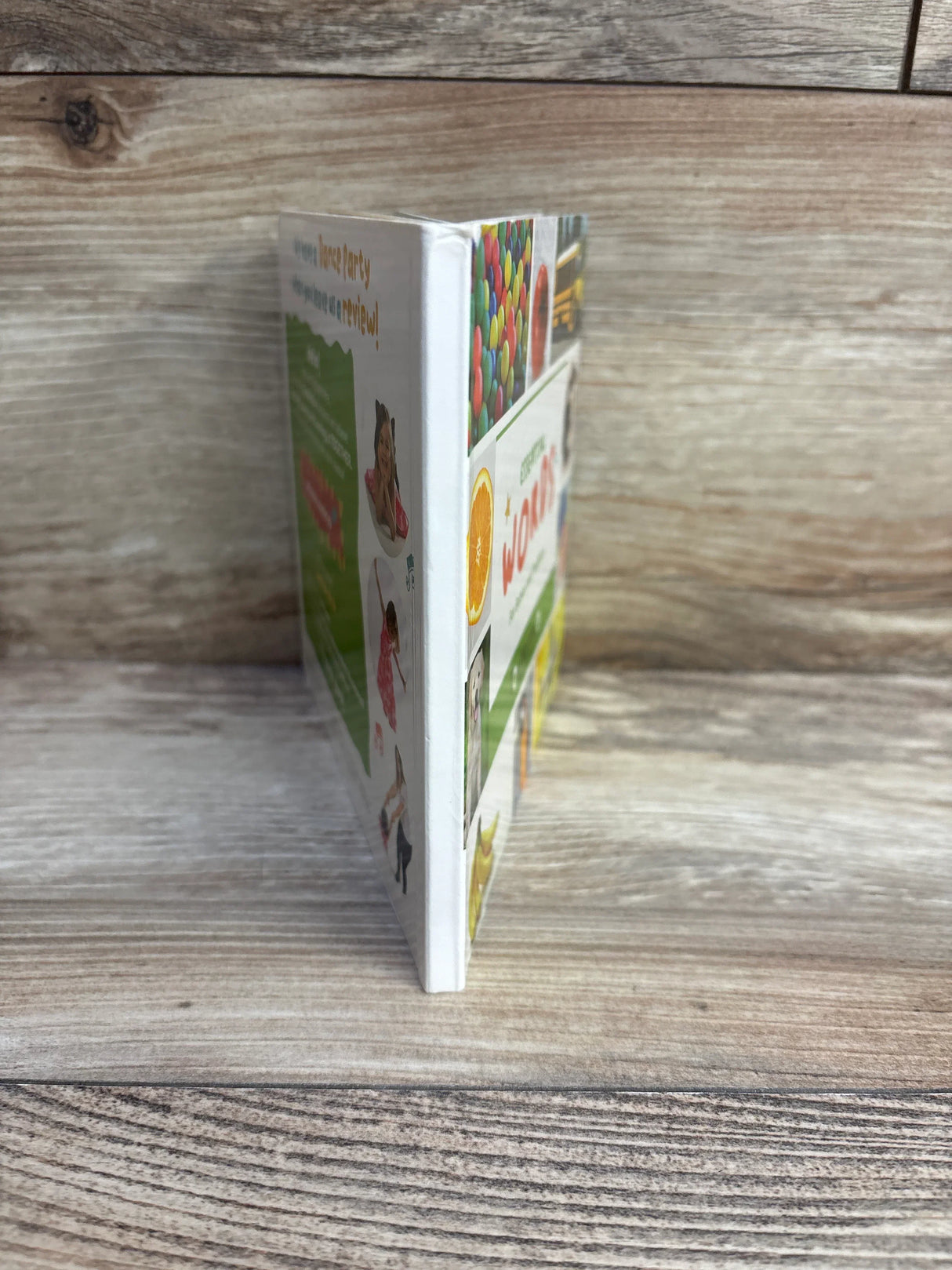 Side view of Essential Words board book showcasing white spine and colorful illustrated cover panels with thick board pages on a wood grain tabletop