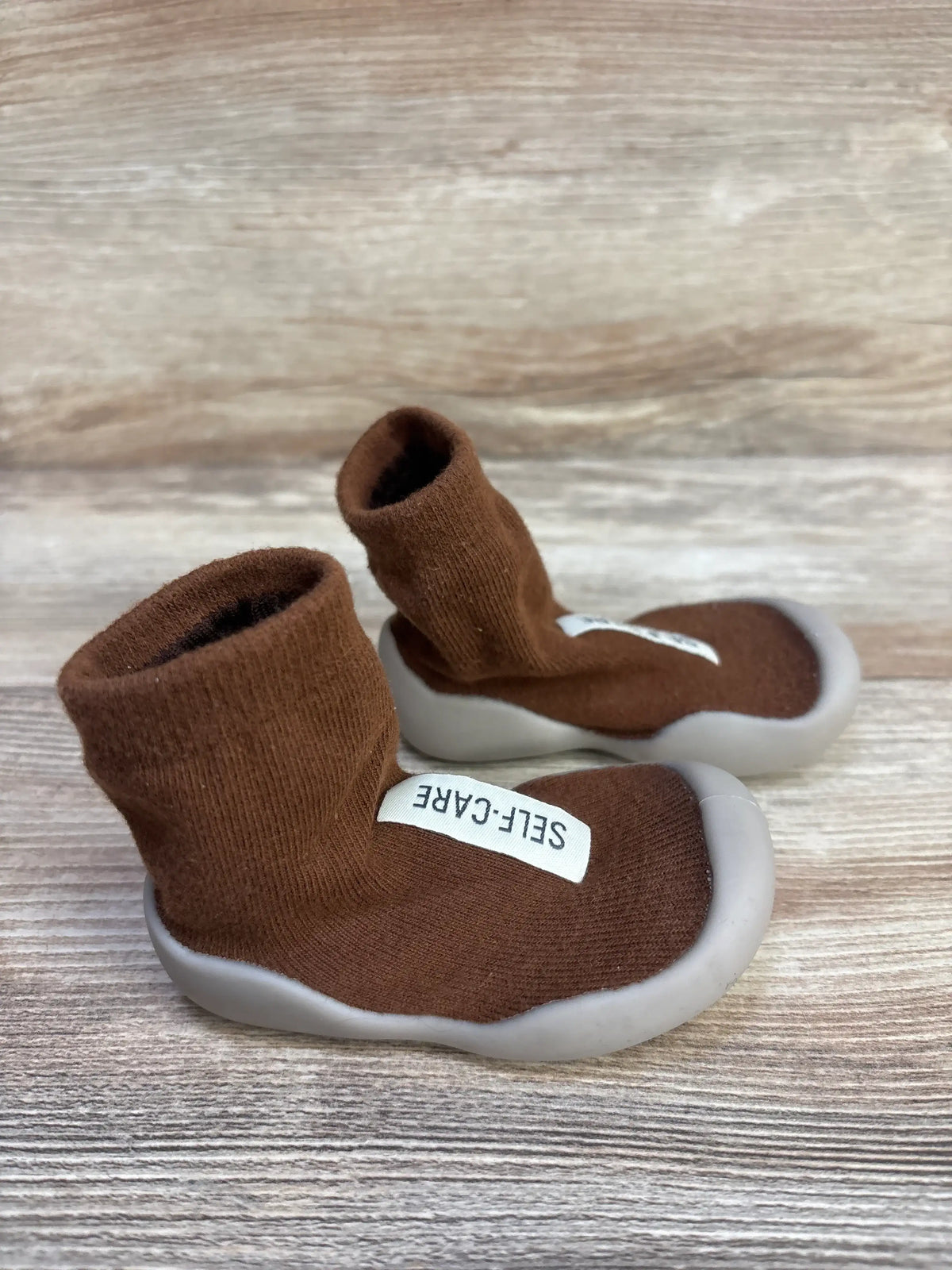 Close view of Tiny Explorings Self-Care Sock Shoes in brown, showcasing sock-like uppers and white soles on a wood-grain surface