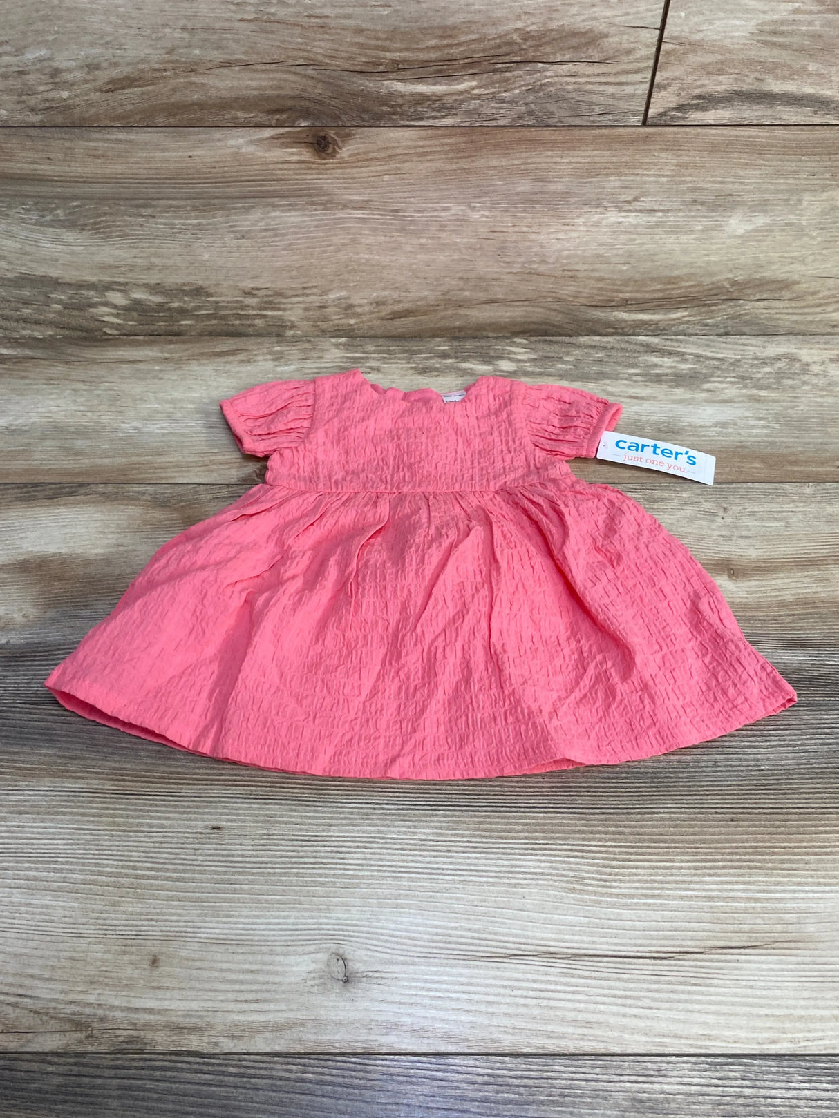 Flat lay of pink baby dress with crinkled texture, puff sleeves, gathered skirt, and Carter's tag on light wood background