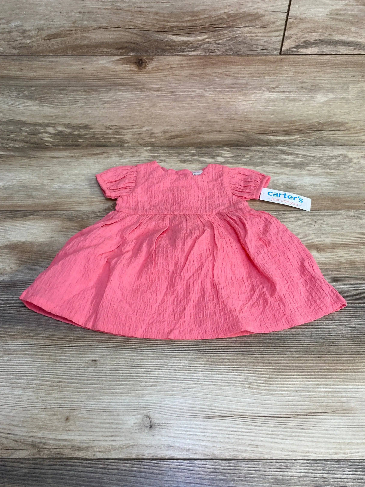 Flat lay of pink baby dress with crinkled texture, puff sleeves, gathered skirt, and Carter's tag on light wood background
