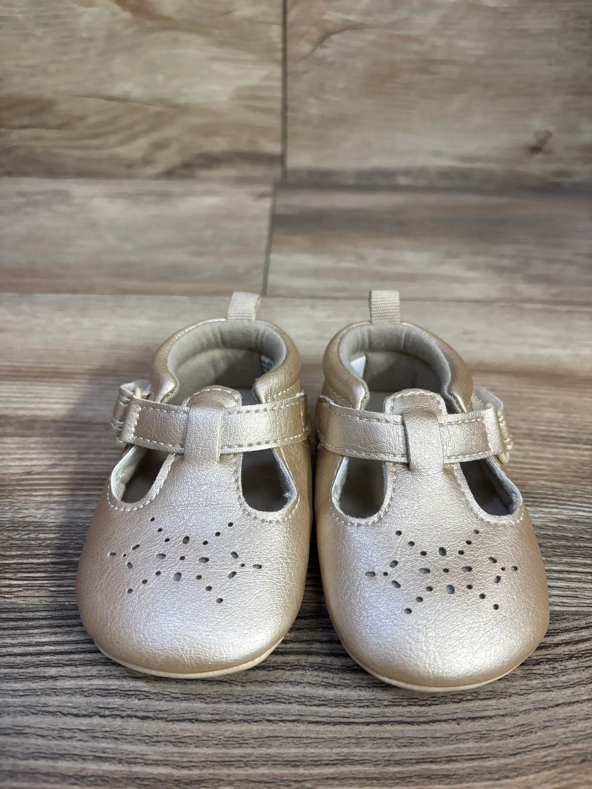 Front view of gold Just One You Stella pre-walker closed-toe sandals with perforations and hook-and-loop straps on wood