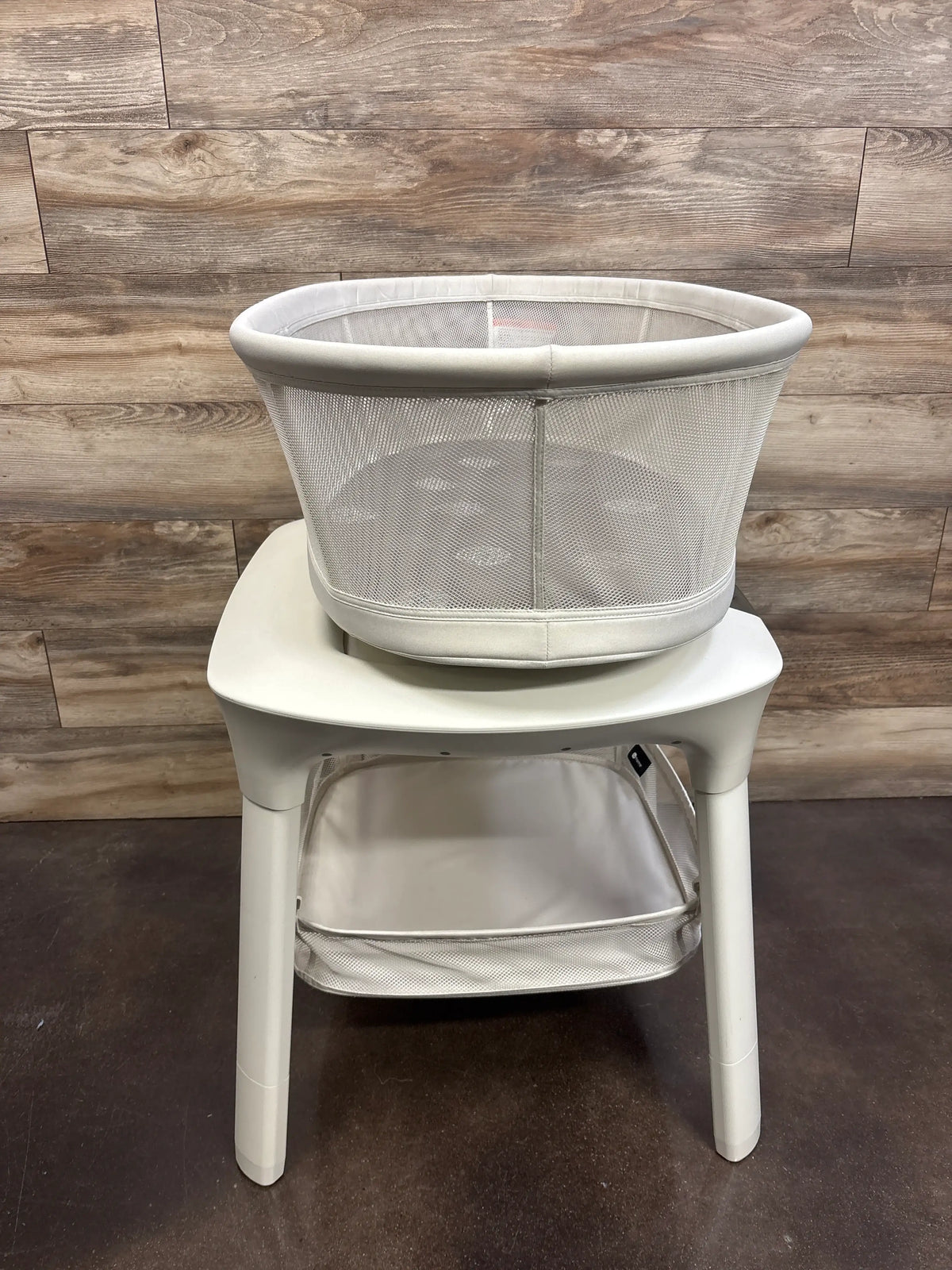 Front view of 4moms mamaRoo Sleep Bassinet showcasing its lower storage basket with a wood-paneled background