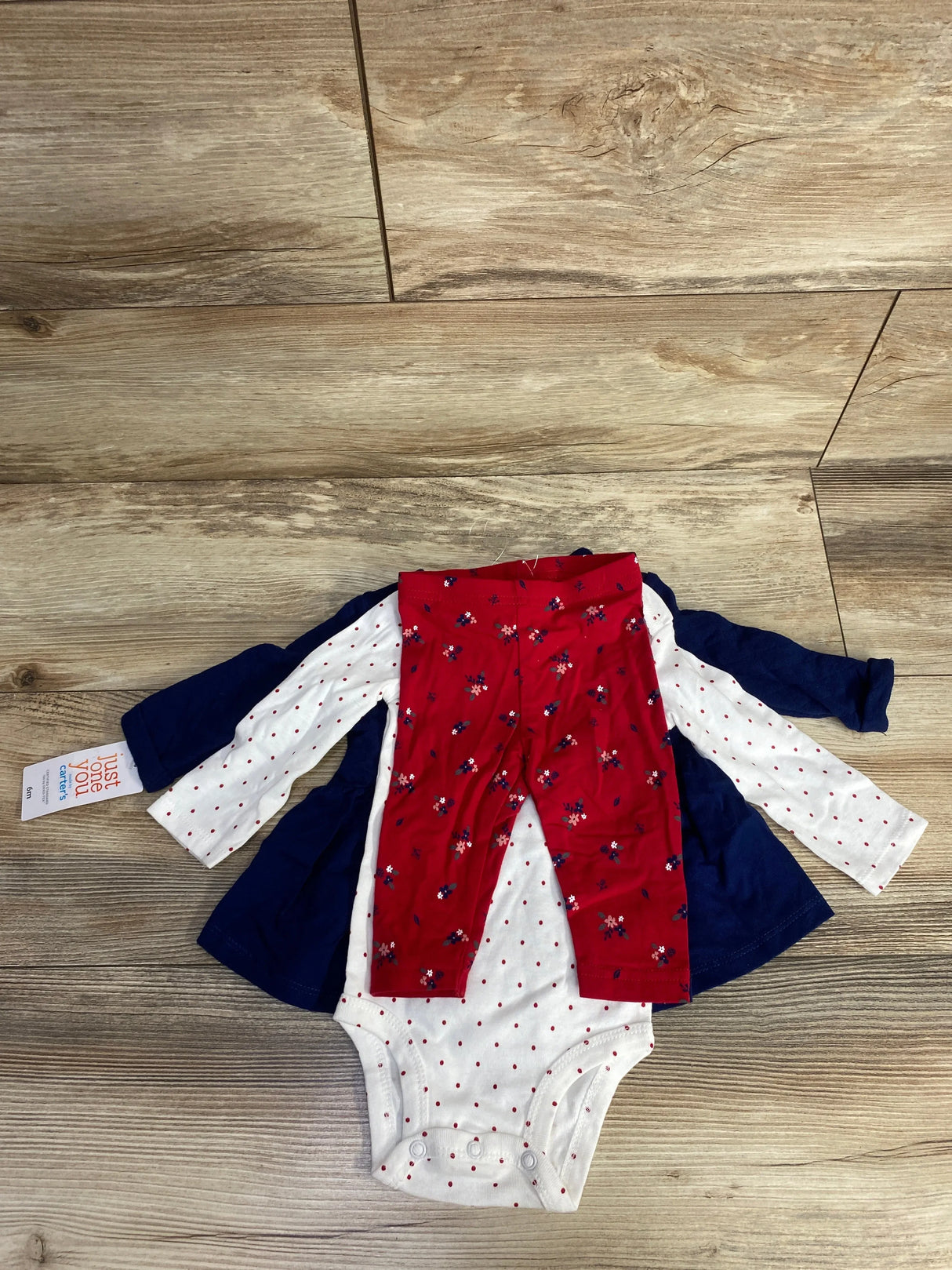 Flat lay of Just One You 3pc set (3-6M) showing navy dress, white polka-dot bodysuit, and red floral leggings on wood floor