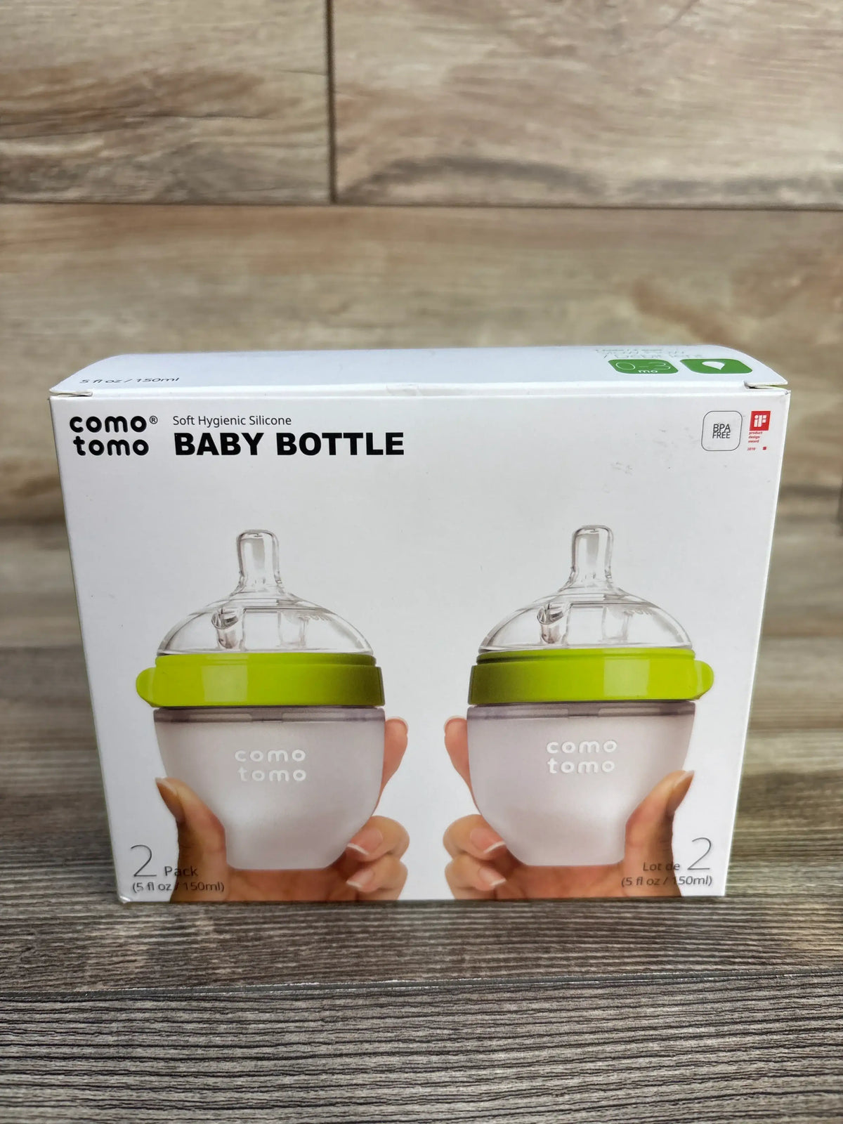 Front view of Comotomo 2-pack baby bottle set showcasing two green-collared silicone bottles in packaging on a wood surface