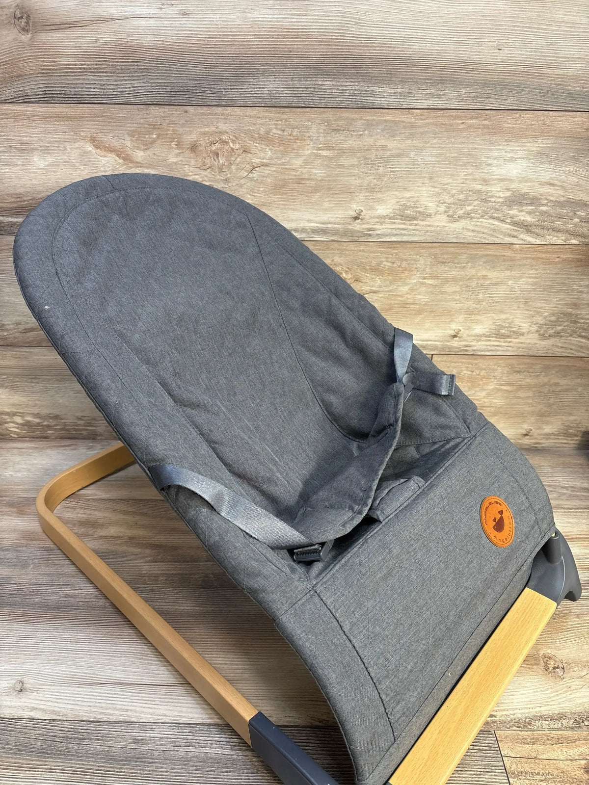 Three-quarter view of ANGELBLISS baby bouncer with grey fabric seat, wooden rocker frame, three-point harness on wood floor