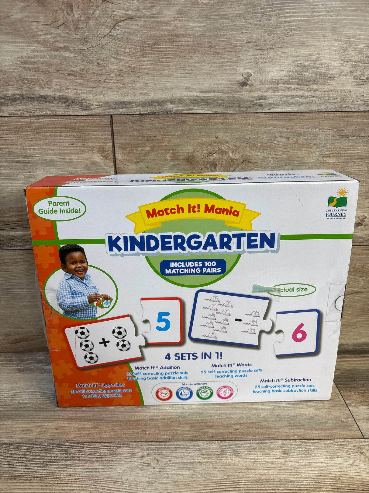 Front view of Match It Mania Kindergarten puzzle box showing Includes 100 matching pairs, sample puzzle-piece cards and child photo on wooden floor