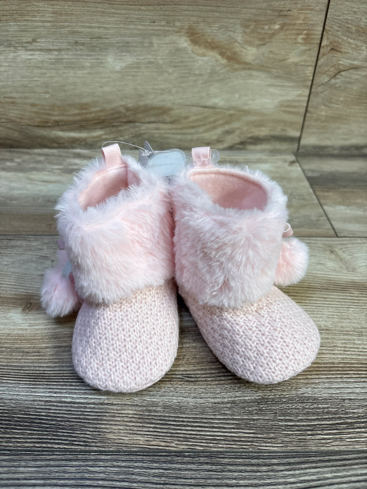 Front view of pink knit baby booties with faux fur cuffs and pom-pom accents on wood background