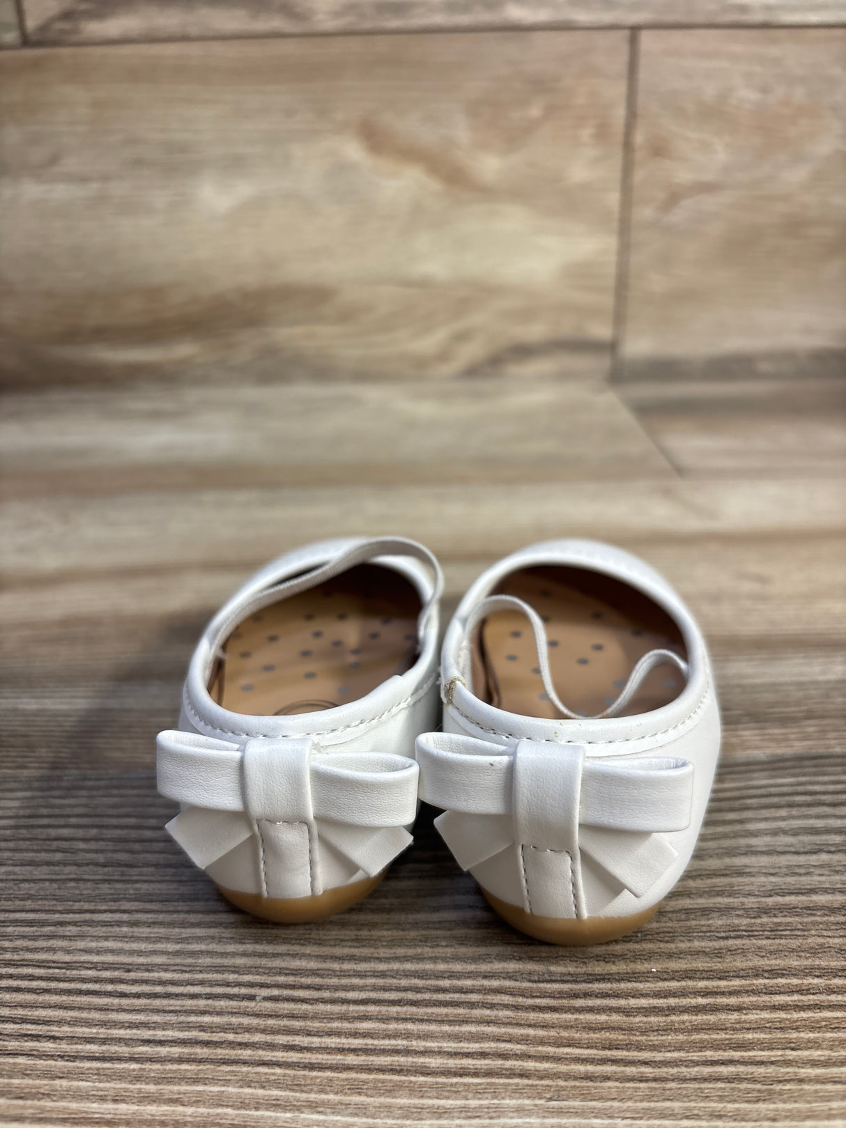 Rear view of white girls' ballet flats (size 2C) showing bow detail on the heels against a woodgrain floor