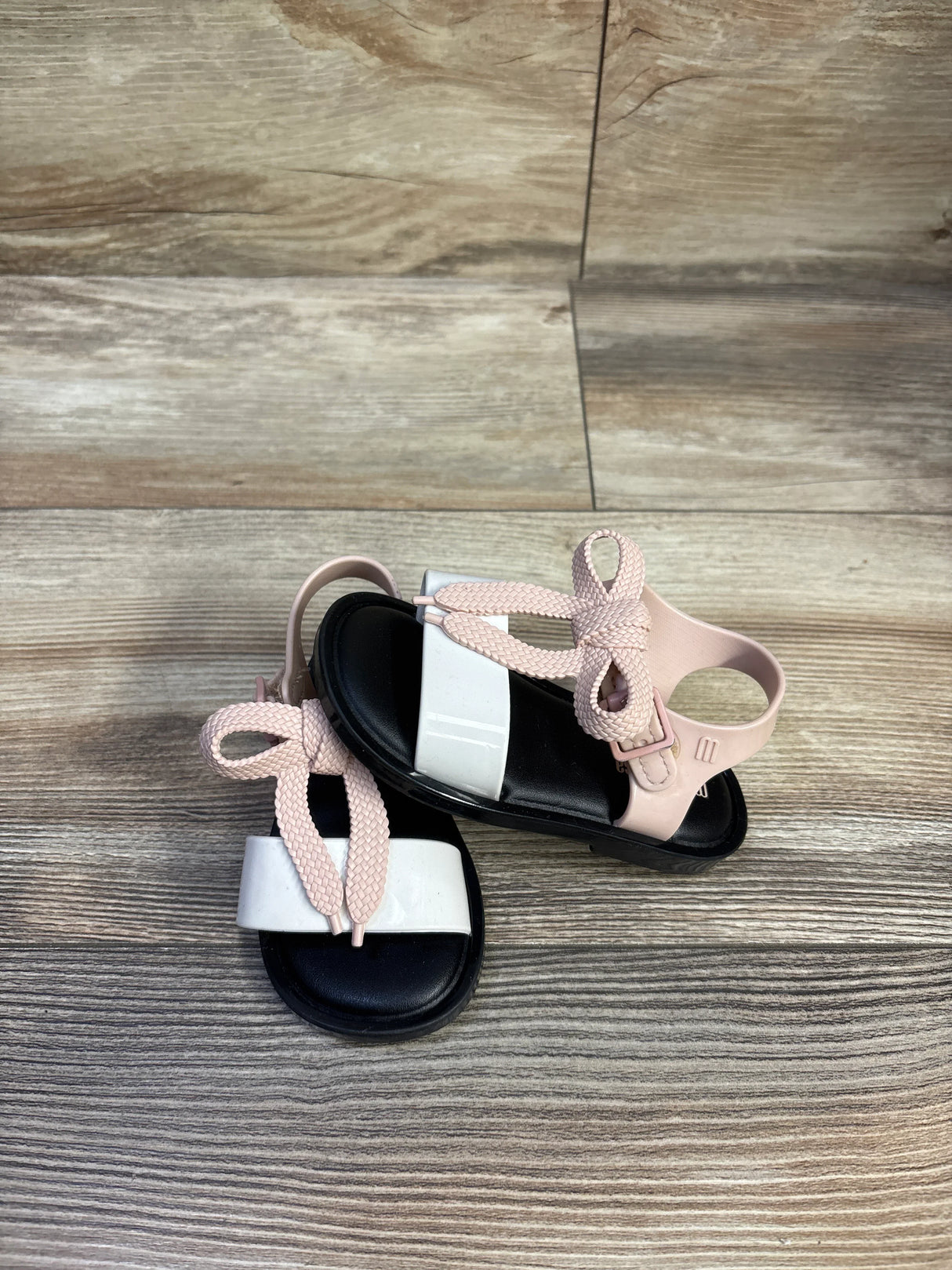 Top-down view of Mini Melissa Mar toddler sandals showcasing black footbed, white glossy toe strap and pink braided ankle ties on wood background