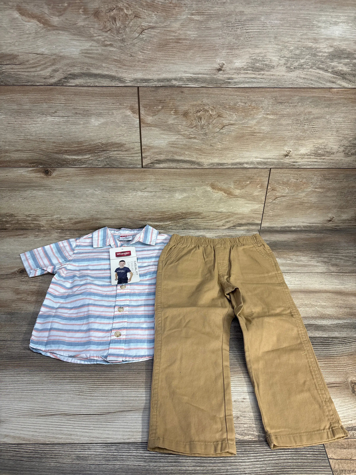 Front view of Wrangler toddler outfit with a striped button-up shirt and tan bottoms on a wood-look background
