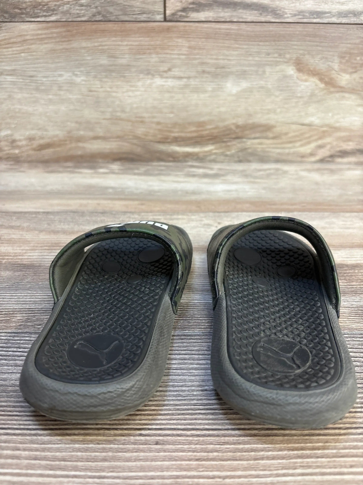 Front view of Puma Cool Cat Camo kids' slides showcasing green camo straps, textured footbeds, and Puma logo on wood floor