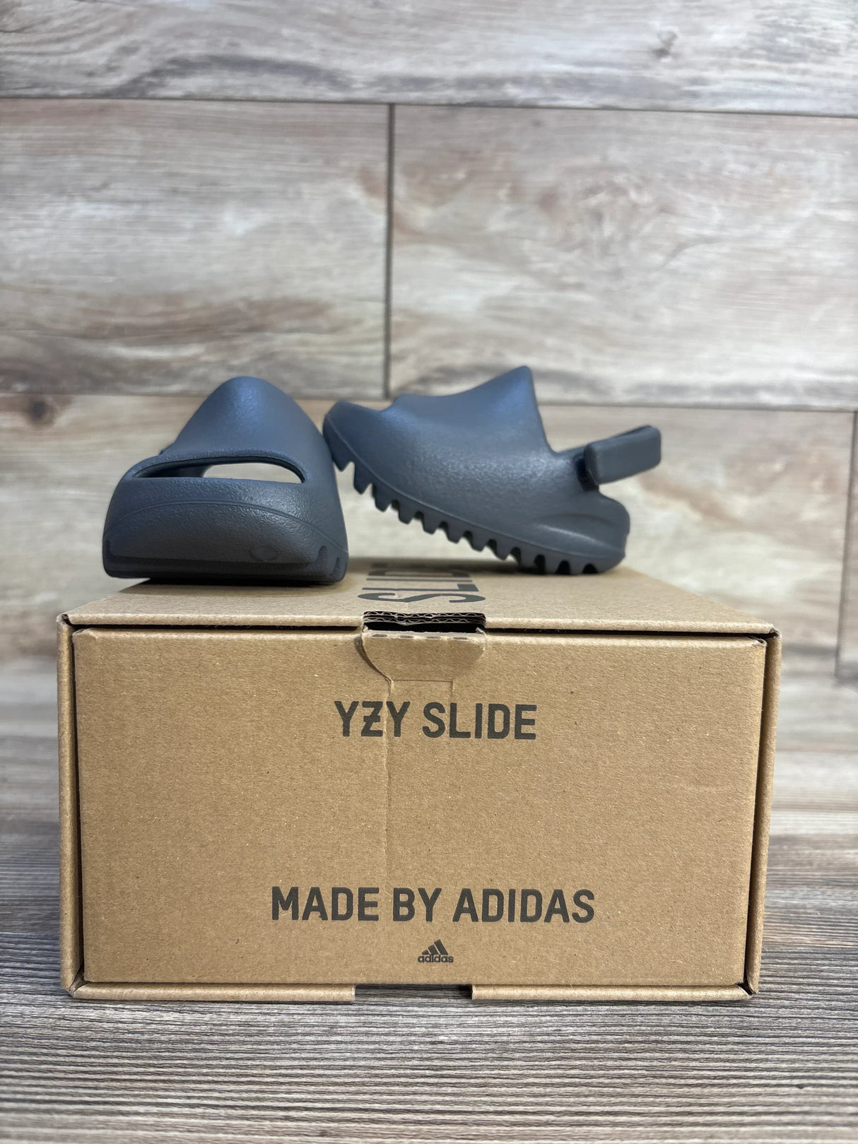 Gray Adidas YZY Slide sandals for kids displayed on cardboard shoe box, wood background