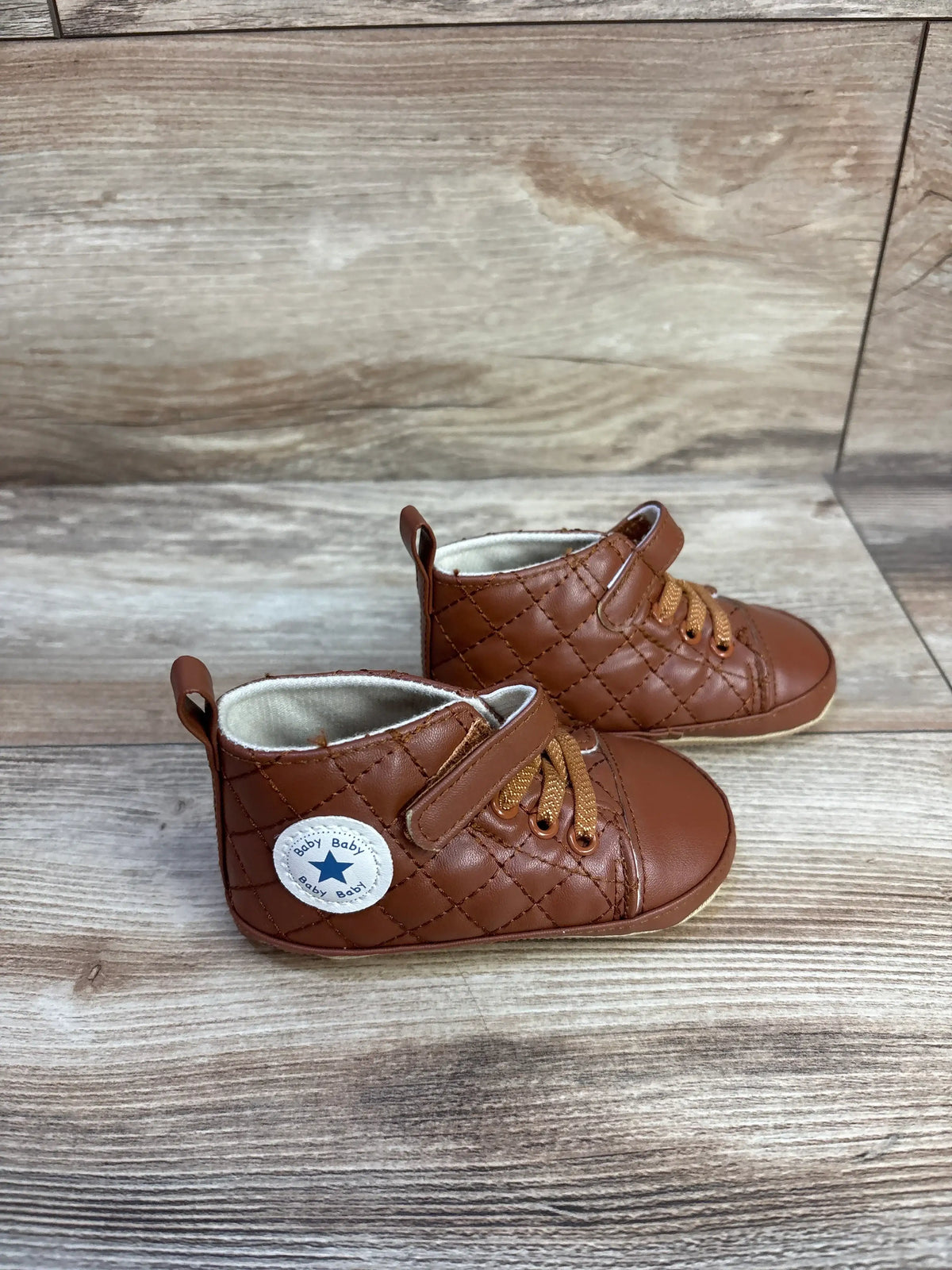 Front and side view of brown faux leather crib shoes showcasing quilted uppers, hook-and-loop straps, and laces on a wood-grain background